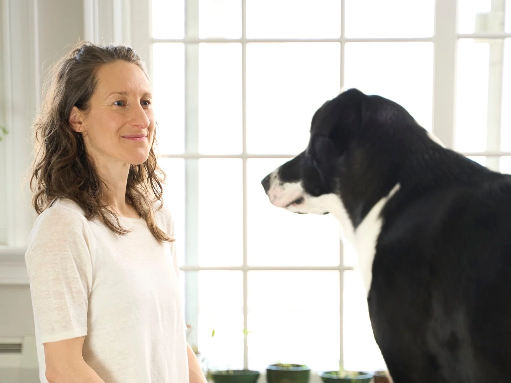 Sharon, a white human with wavy brown locks and cream top gazes at Muggins, a large black-and-white floppy-eared dog. The two hold their breath in a frozen moment.