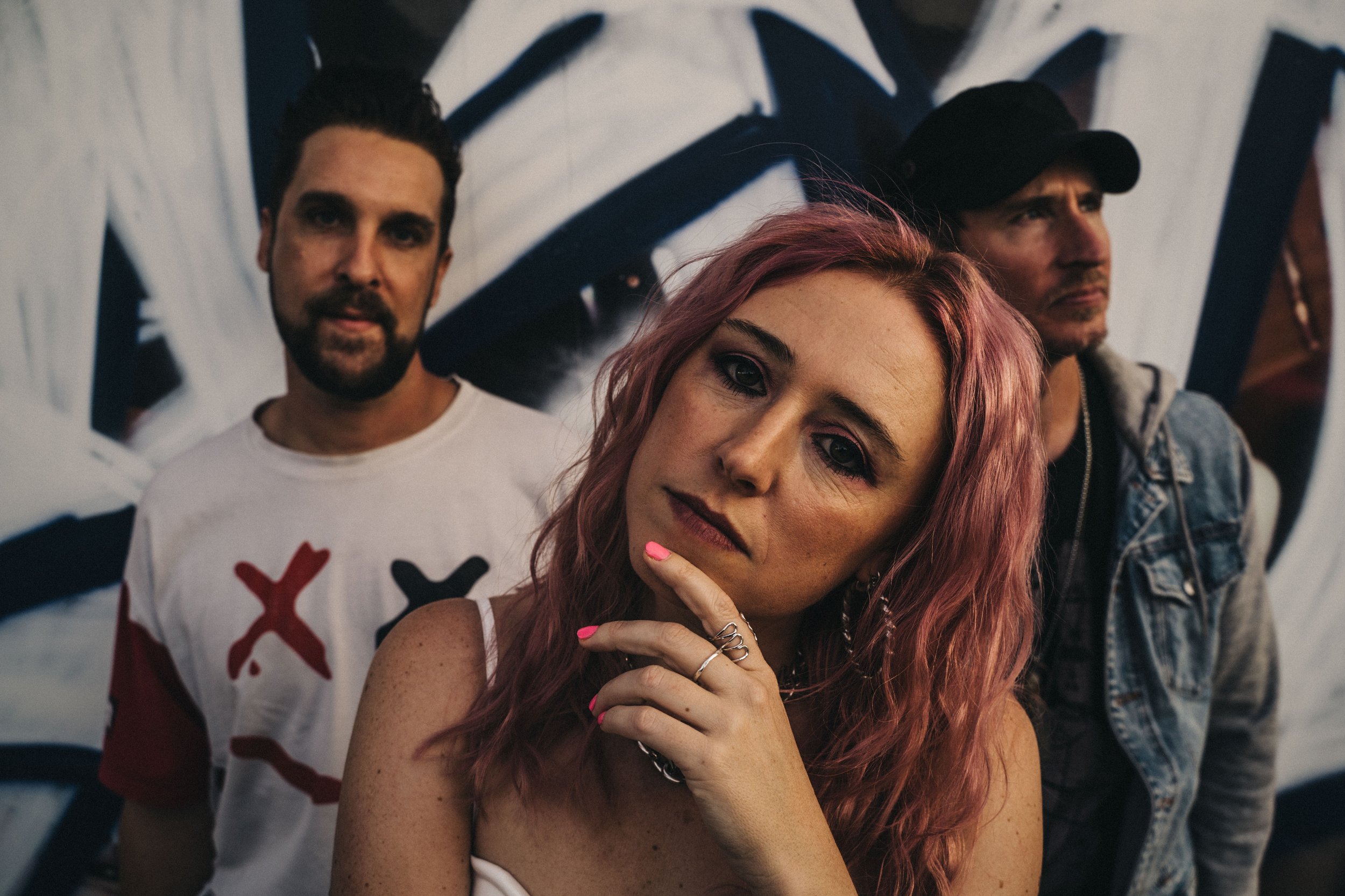 Three people standing in front of a graffiti wall, with a woman in the foreground holding her chin, a man in the middle wearing a white shirt with red and black designs, and another man in a denim jacket and black cap in the background.
