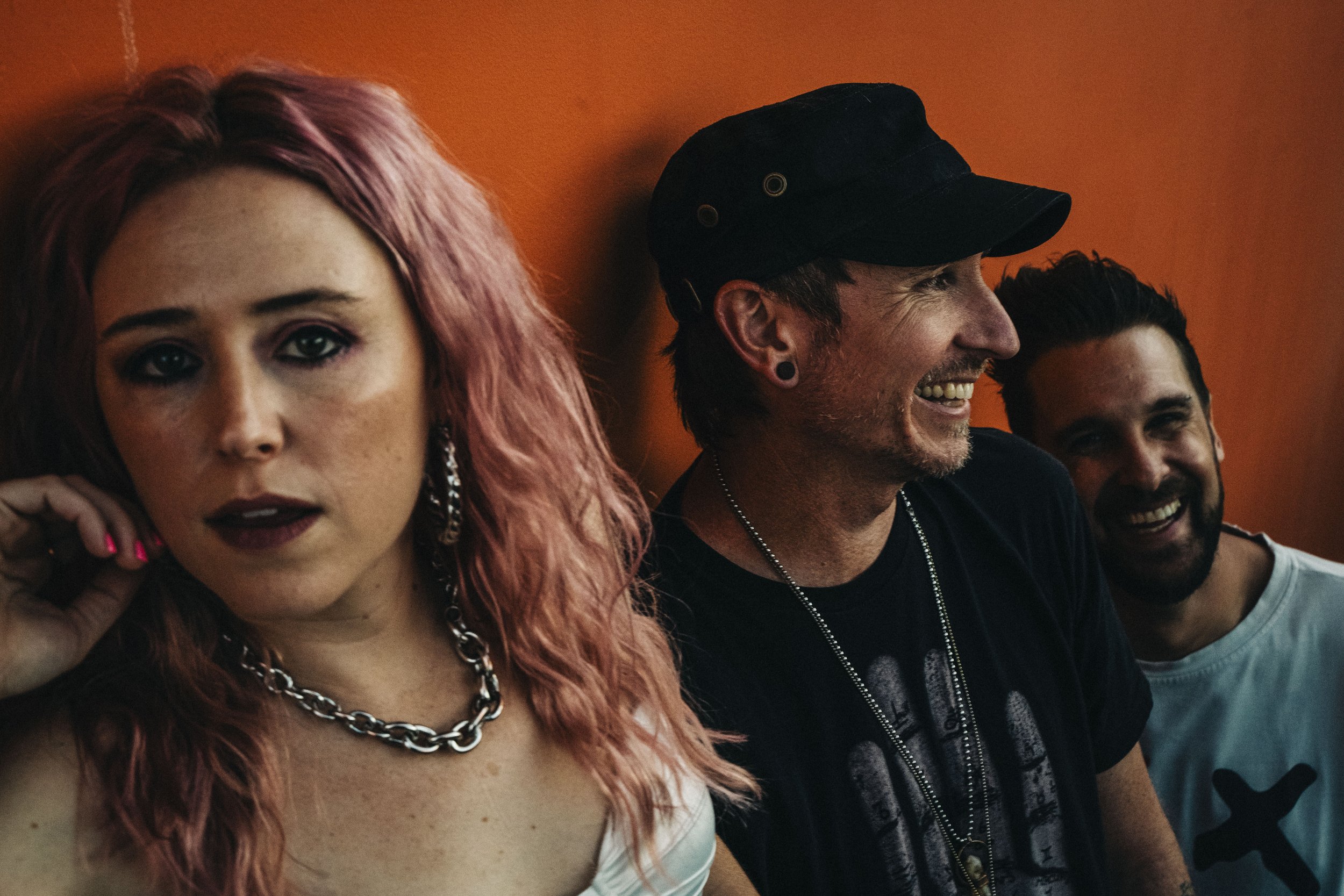 Three people standing against an orange wall, smiling. One person has pink hair and is wearing a chain necklace; the other two have dark hair, one wearing a cap, all appear to be in a relaxed and joyful mood.
