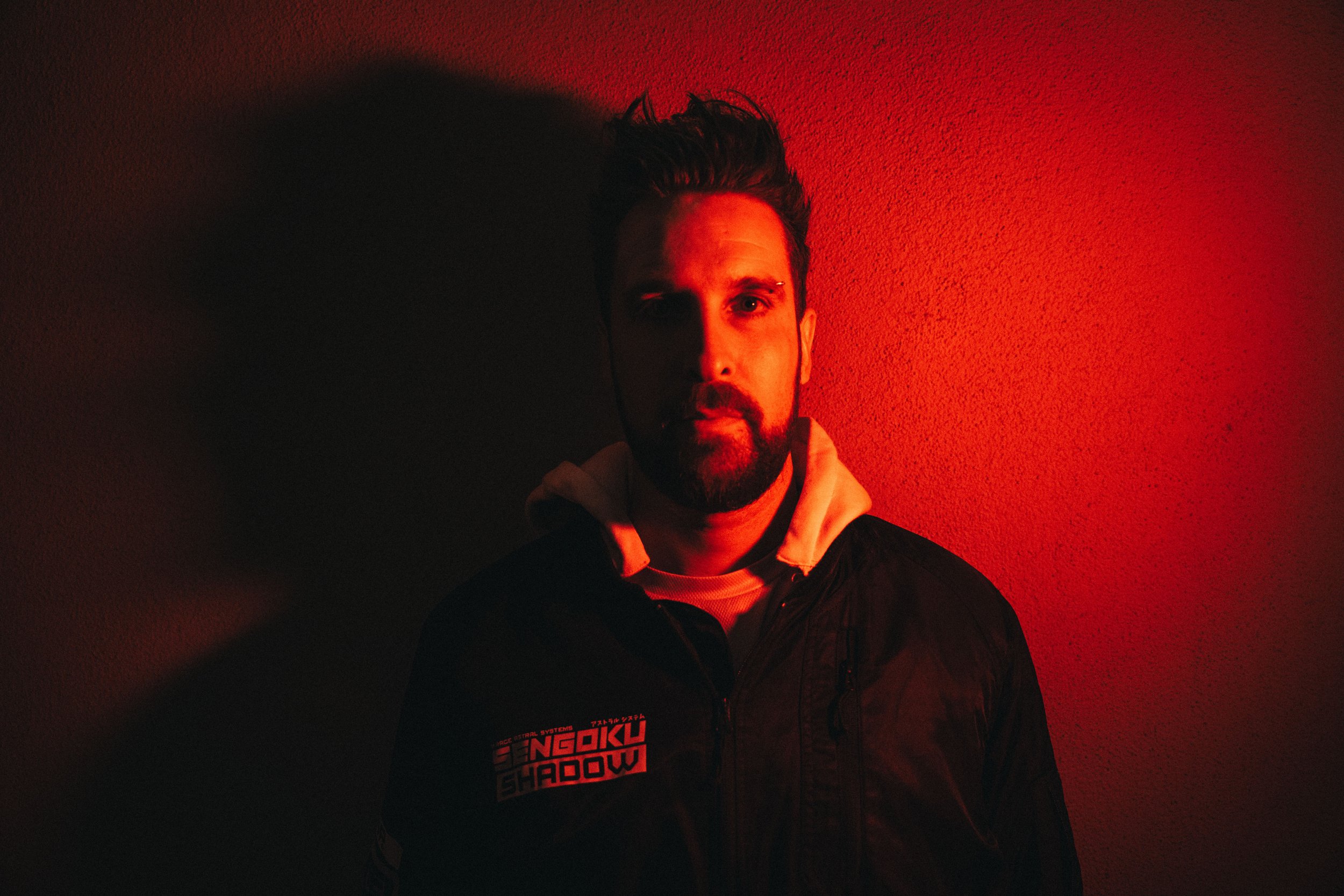 Man with beard and mustache in a jacket, standing against a red-lit wall.