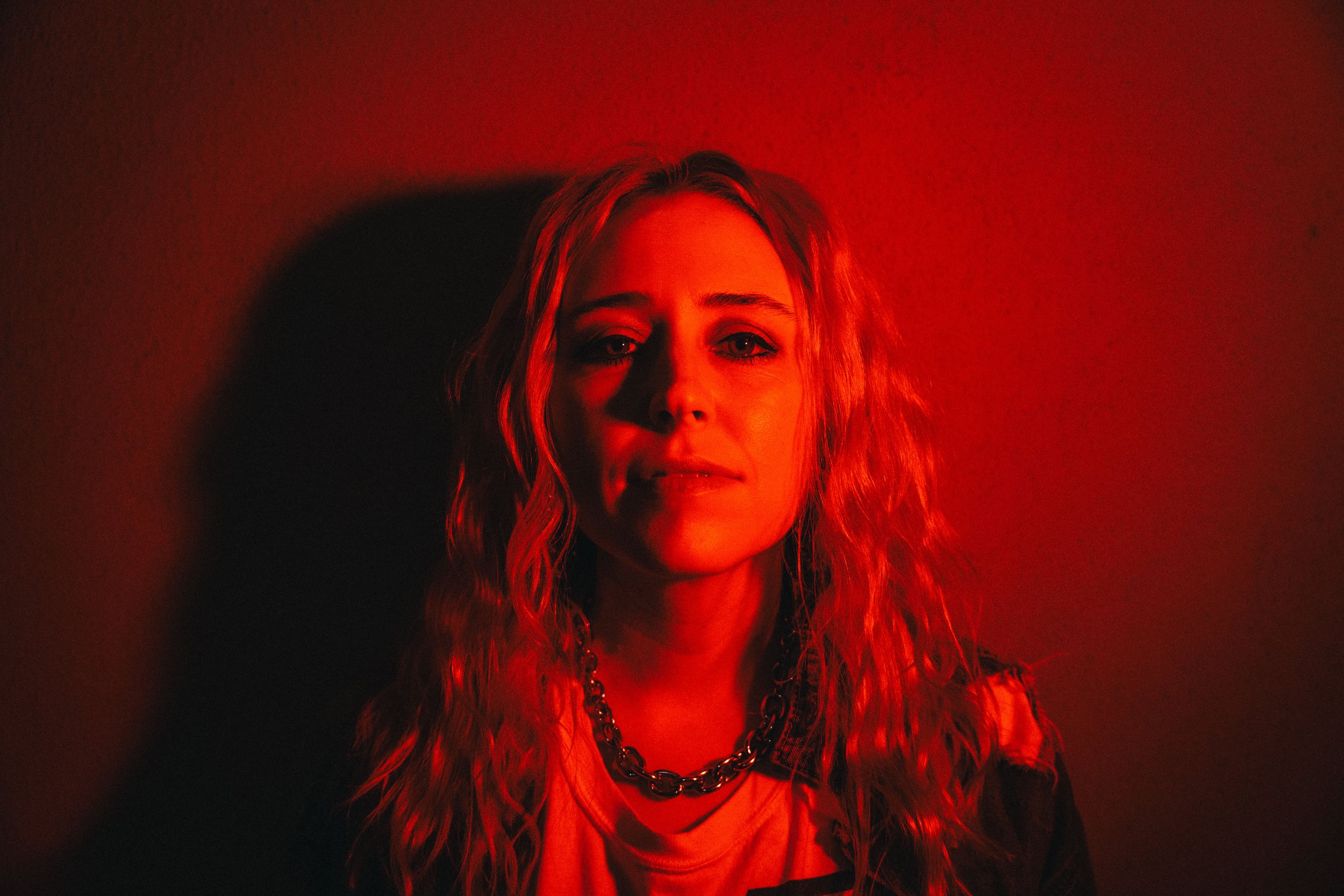 Person with long, wavy hair lit by red light, wearing a necklace, standing against a red wall.