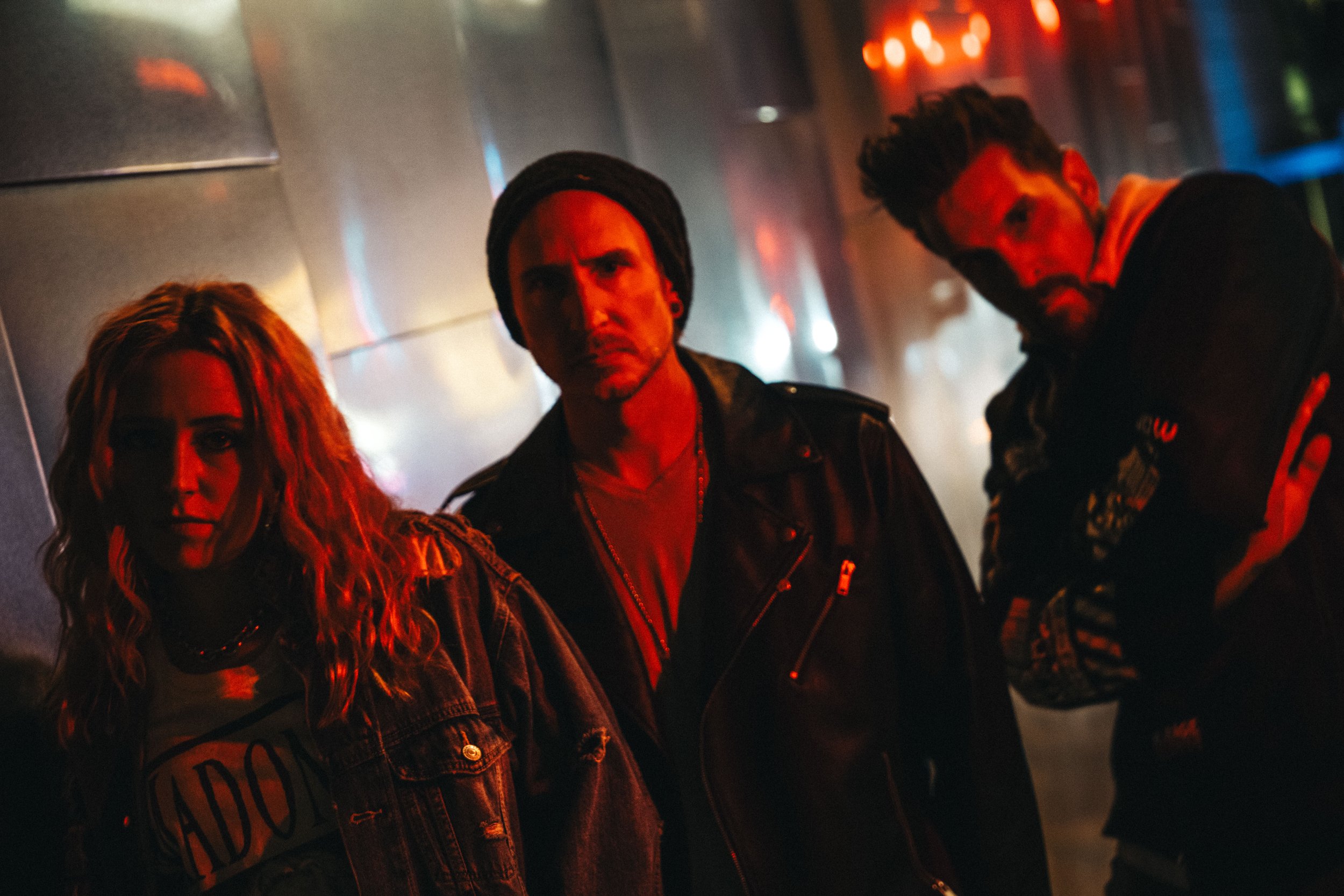 Three people standing in a dimly lit urban setting, featuring red and blue lighting. The person on the left has long blonde hair, the middle one wears a beanie and a leather jacket, and the right one has a beard and crosses their arms. The background