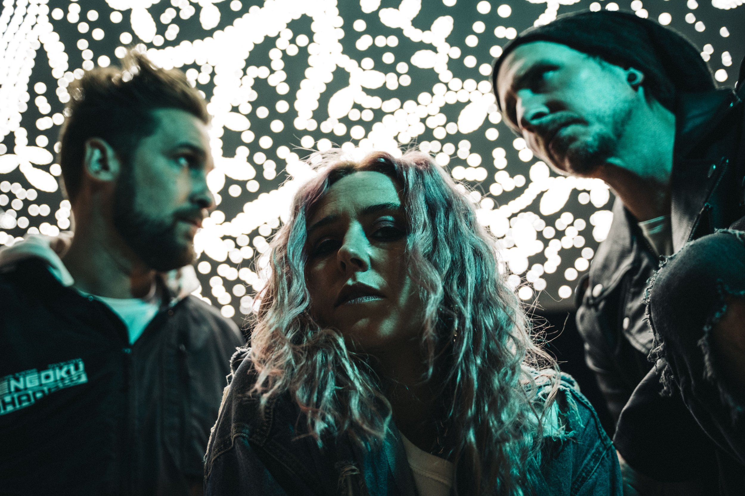 Three people in front of a bright, abstract background with lights, looking serious; two men and one woman with wavy hair.