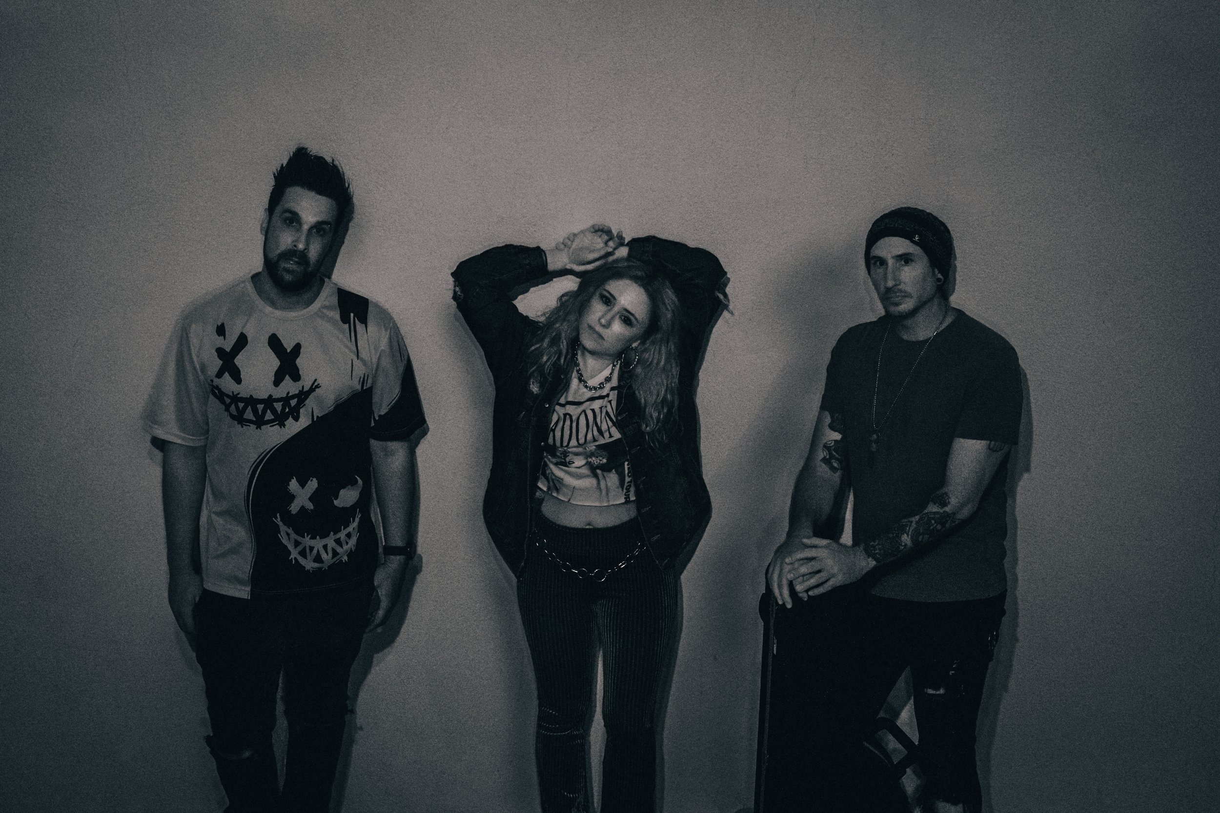 Black and white photo of three people posing against a wall. The person on the left wears a graphic T-shirt, the central person wears a jacket and pants, and the person on the right has a beanie and tattoos.