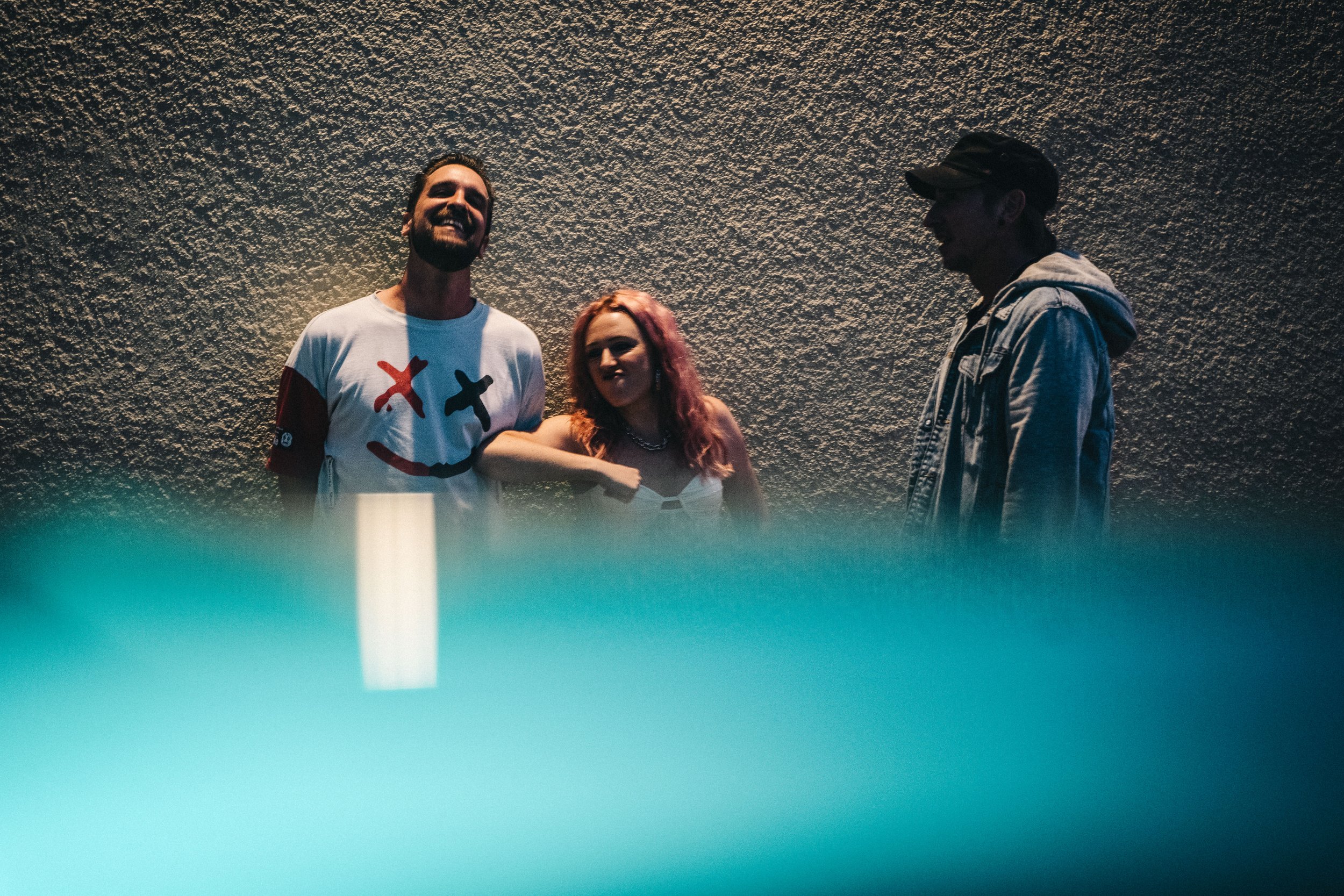 Three people standing against a textured wall, smiling and talking. The foreground is blurred with a teal hue.