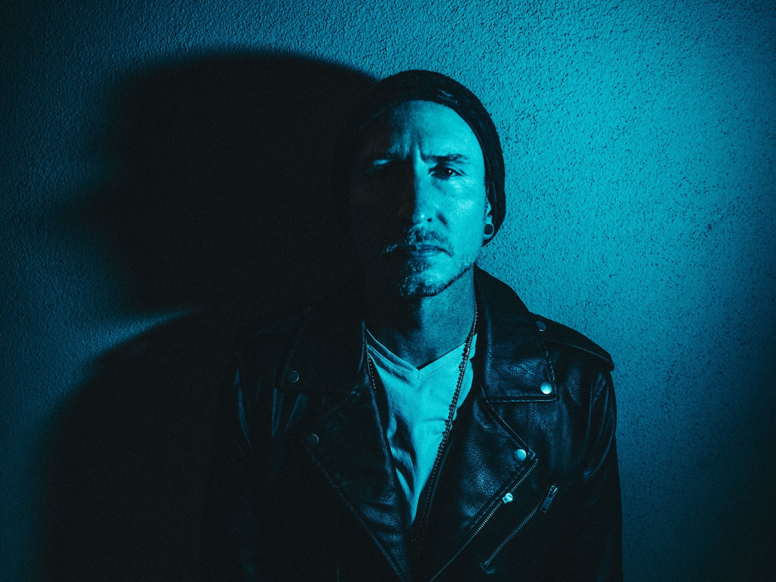 Man wearing leather jacket and beanie in blue lighting against a textured wall