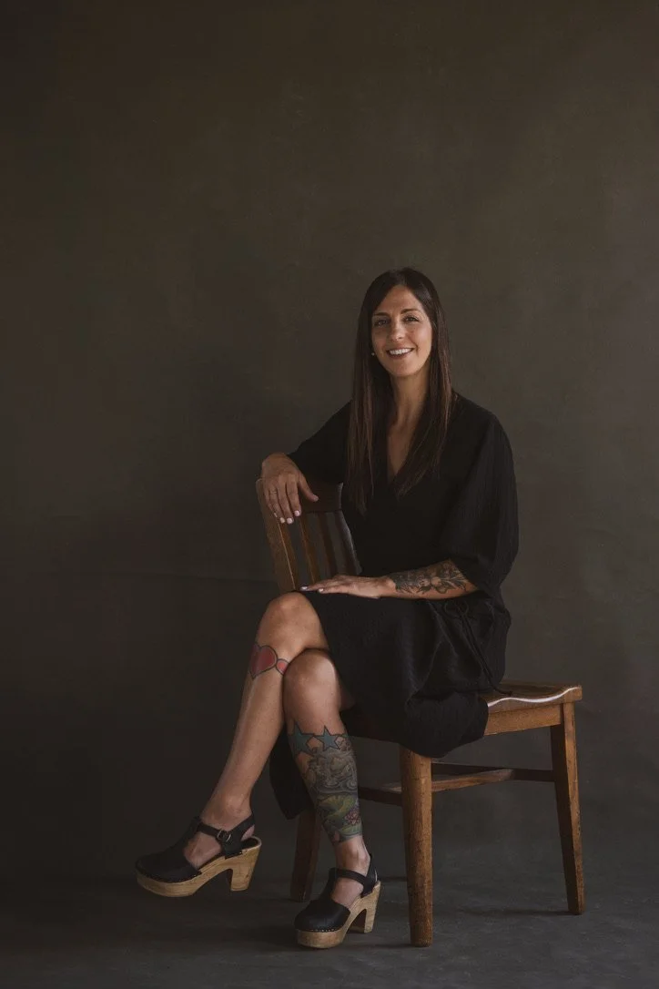 A woman with long dark hair and tattoos on her legs and arm, sitting on a wooden chair against a dark background, wearing a black dress and black platform shoes, smiling at the camera.
