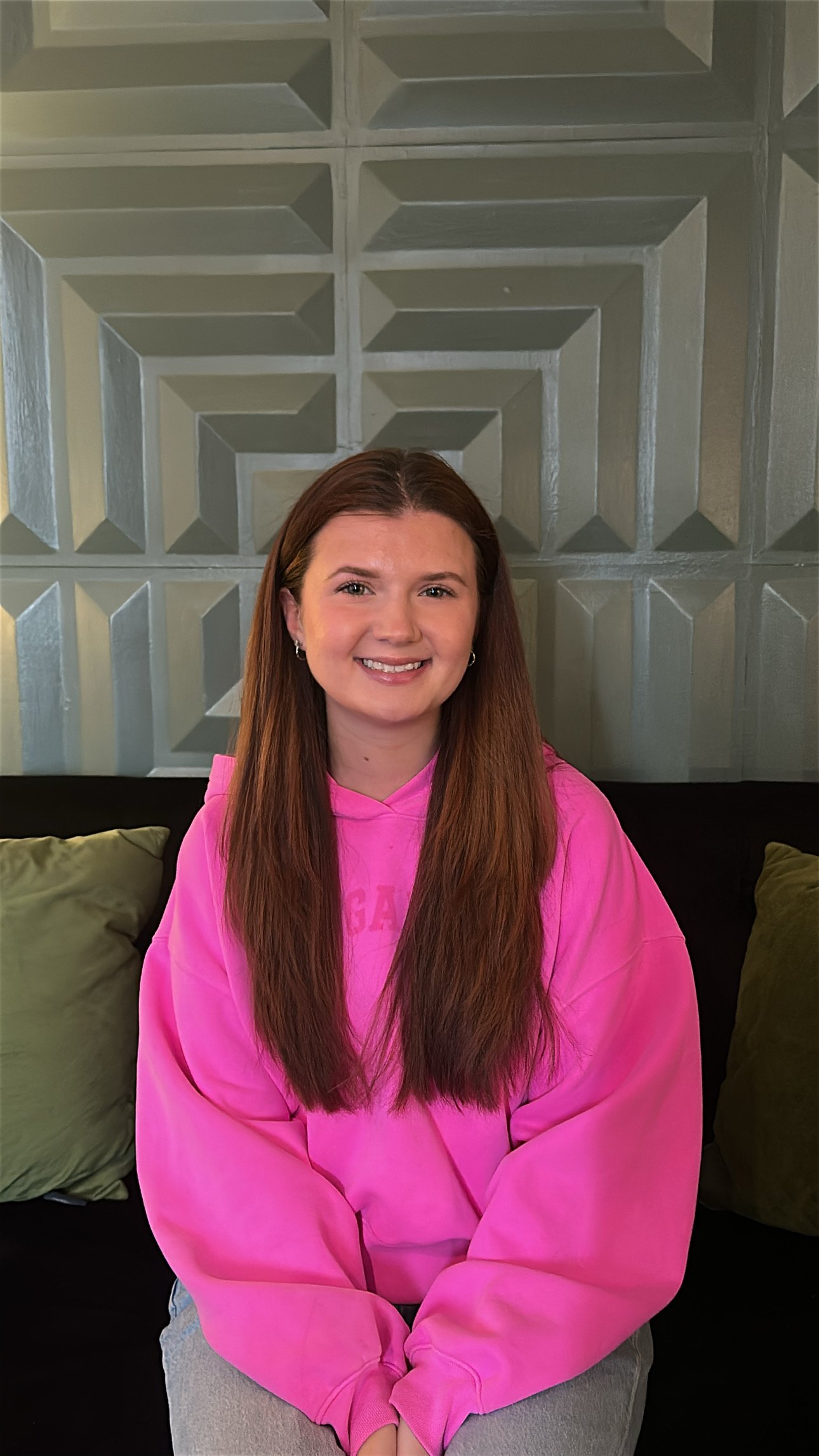 A woman with red hair taking a selfie inside a home, smiling and wearing a light blue sweater with buttons.