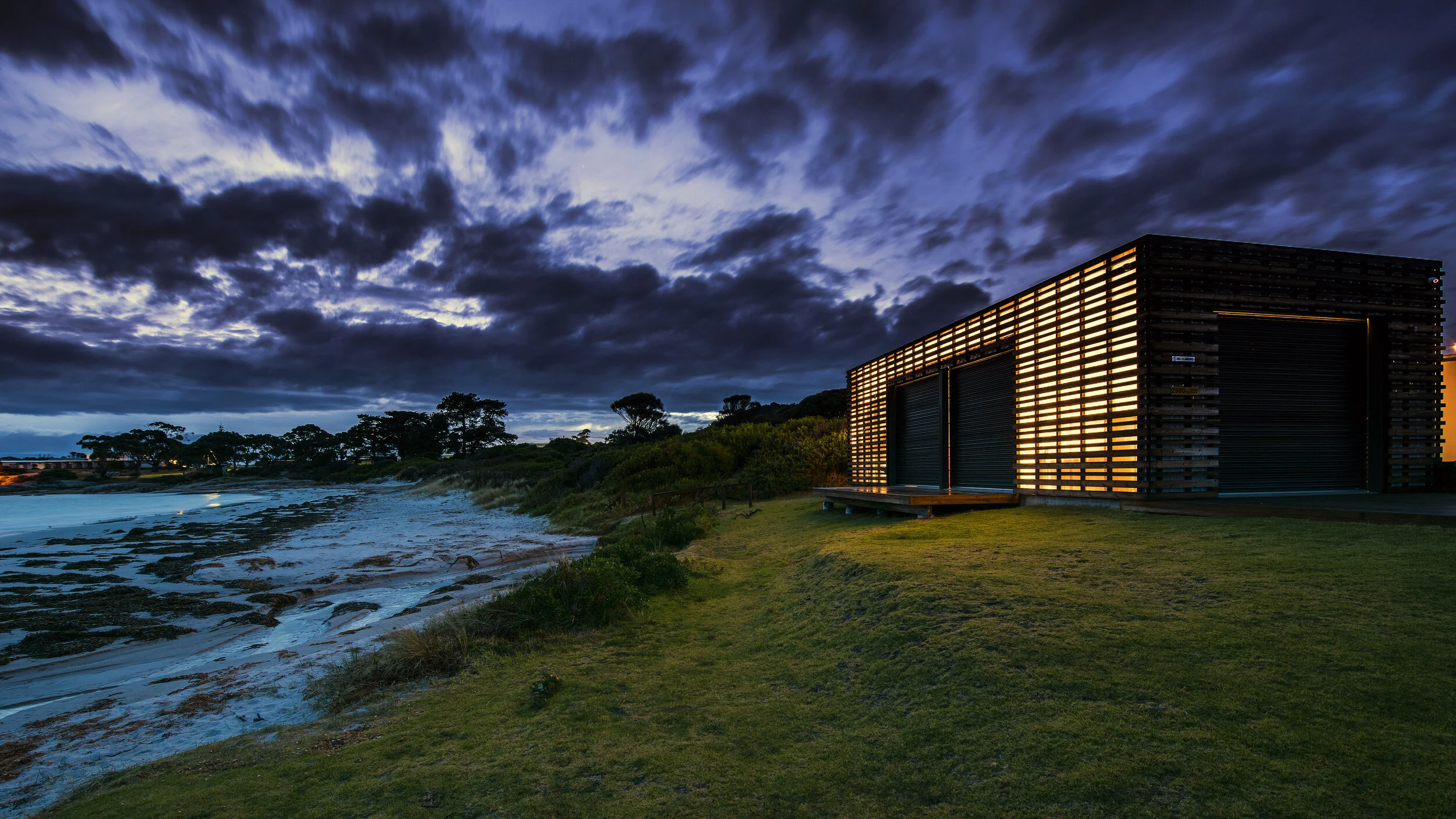 A modern building with horizontal wooden slats, illuminated from within, located on a grassy area near the shoreline during dusk with a cloudy sky overhead.
