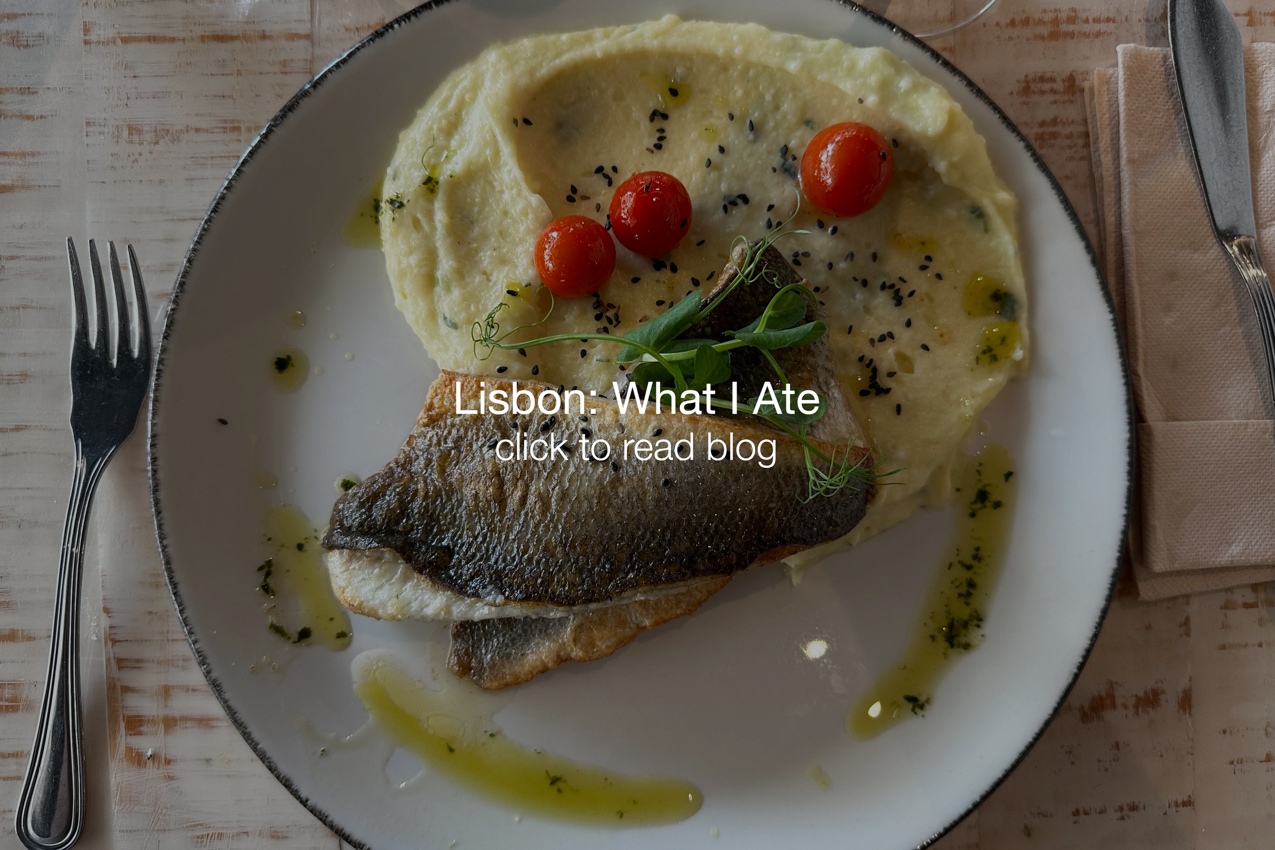 A plate of fish with crispy skin, mashed potatoes, cherry tomatoes, microgreens, black sesame seeds, and green herb oil drizzle, placed on a rustic wooden table with a fork on the left and a knife on the right.