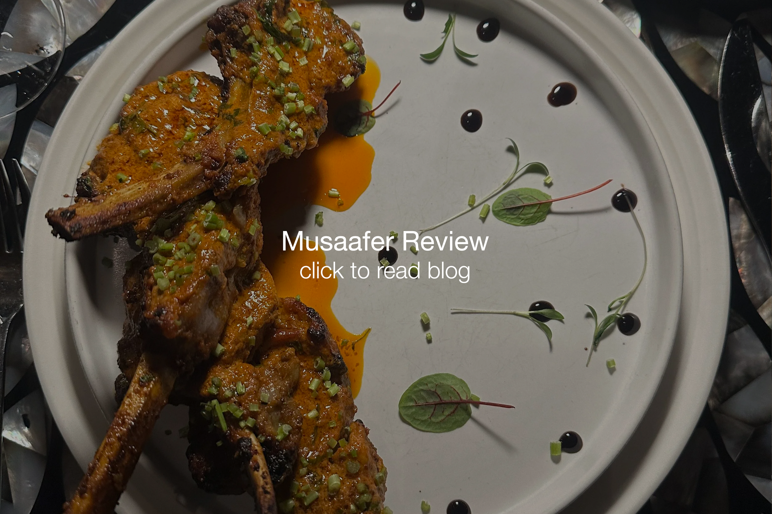 A white plate with grilled meat chops topped with herbs and spices, drizzled with sauce, garnished with chopped green onions and microgreens, decorated with black and green sauce dots and microgreens.