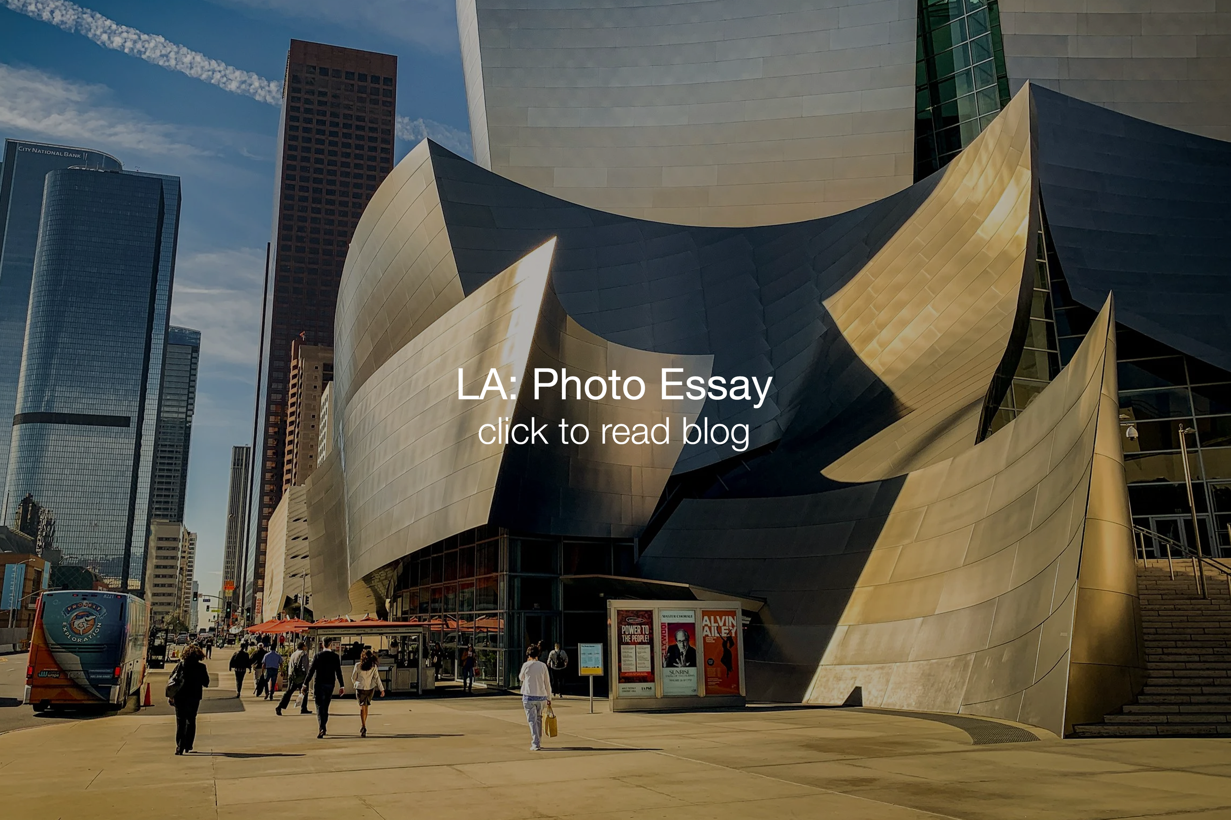 People walking on a city sidewalk in front of the Walt Disney Concert Hall in Los Angeles, with tall skyscrapers in the background on a clear day.