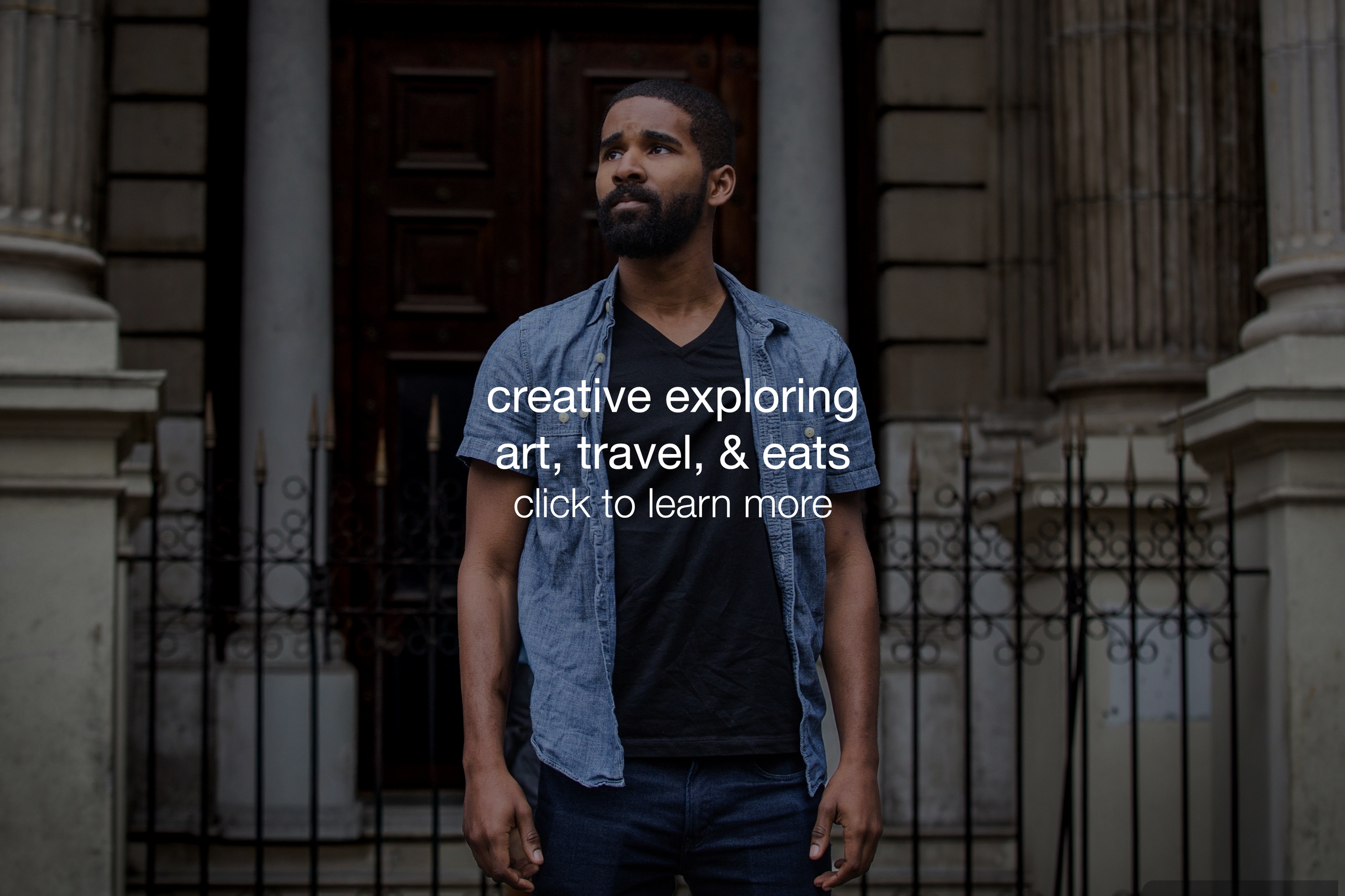 A young man with a beard and short hair standing in front of a gated building entrance, wearing a denim shirt over a black t-shirt, looking off to the side.