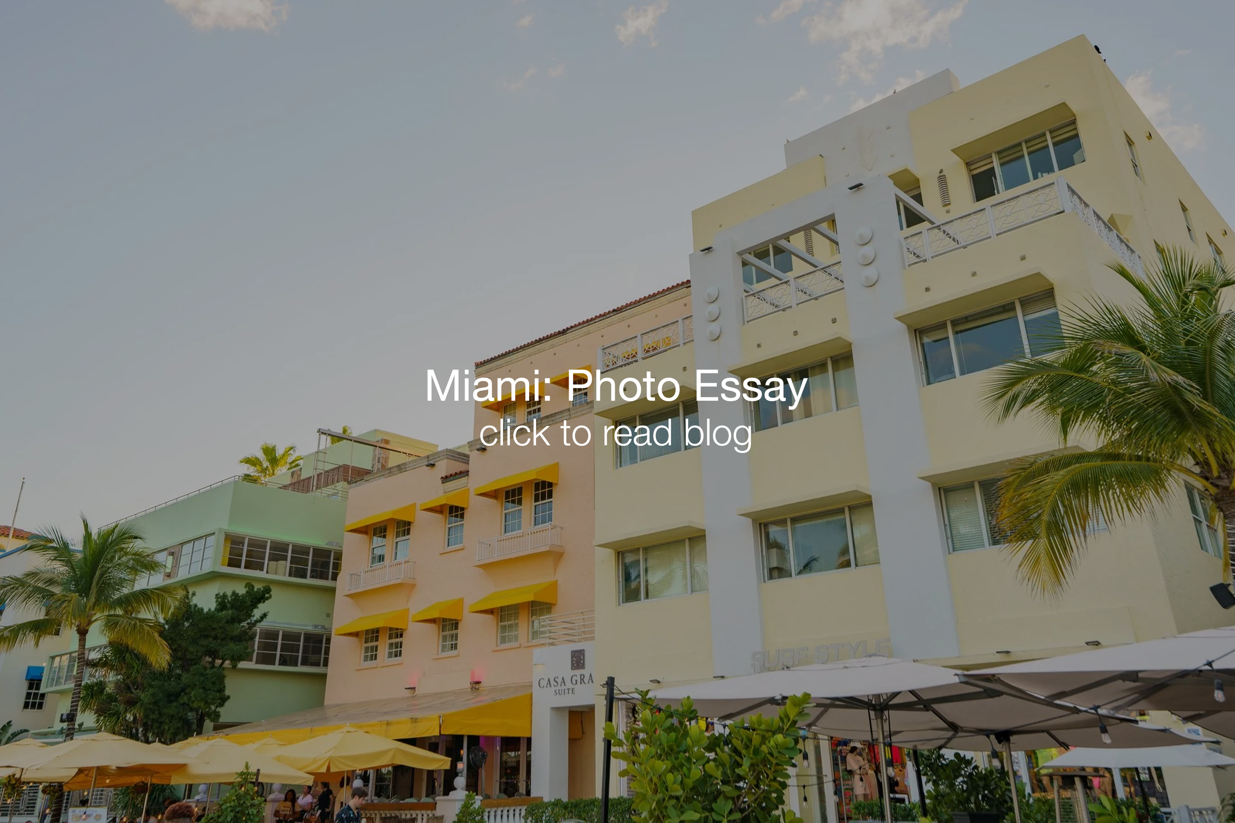 Colorful multi-story buildings in Miami with balconies, palm trees, outdoor seating with umbrellas, and people dining outside.