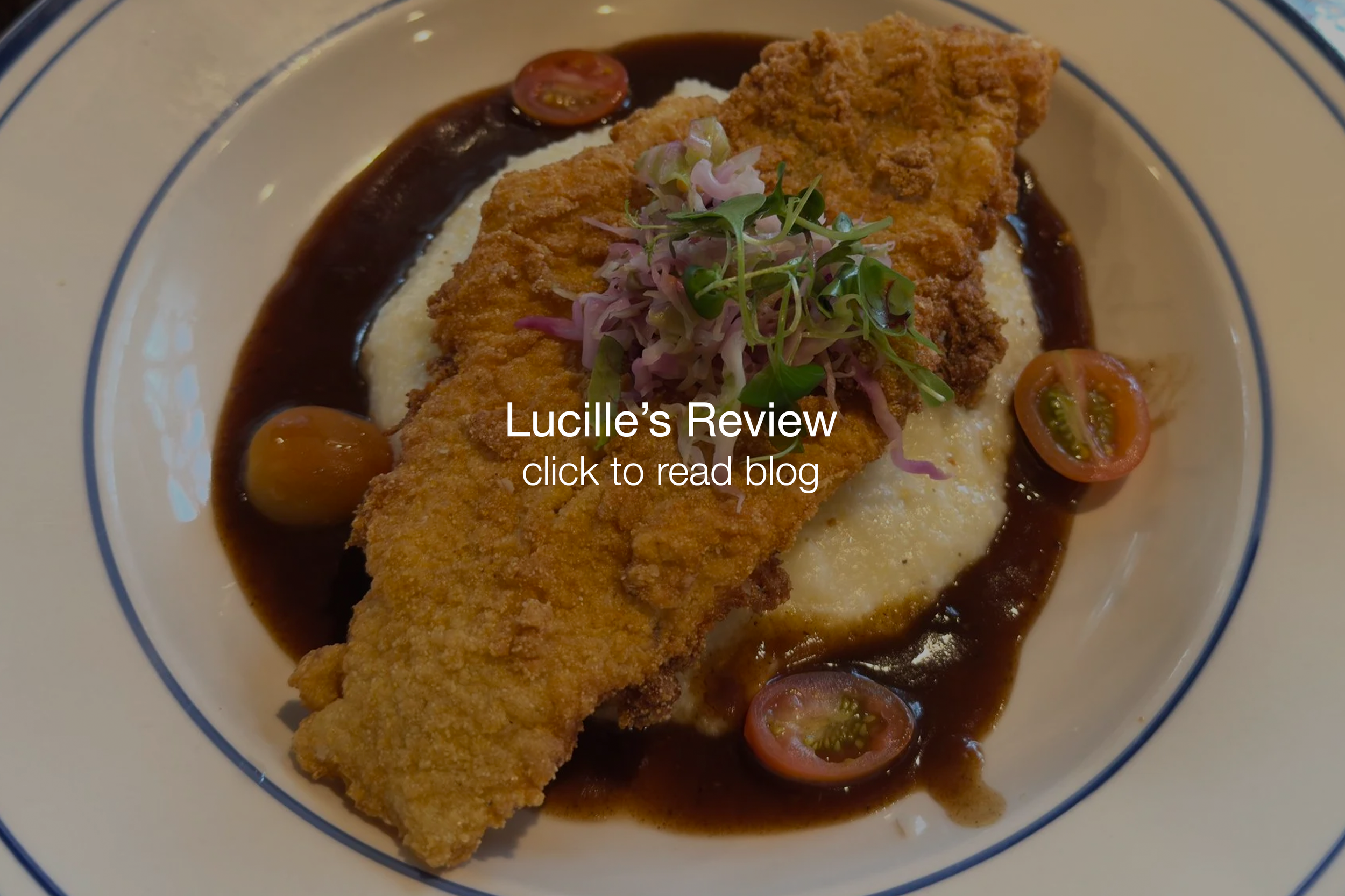 A breaded fried fish fillet topped with microgreens on a bed of mashed potatoes, garnished with cherry tomato halves, and drizzled with dark brown gravy on a white plate with blue rim.