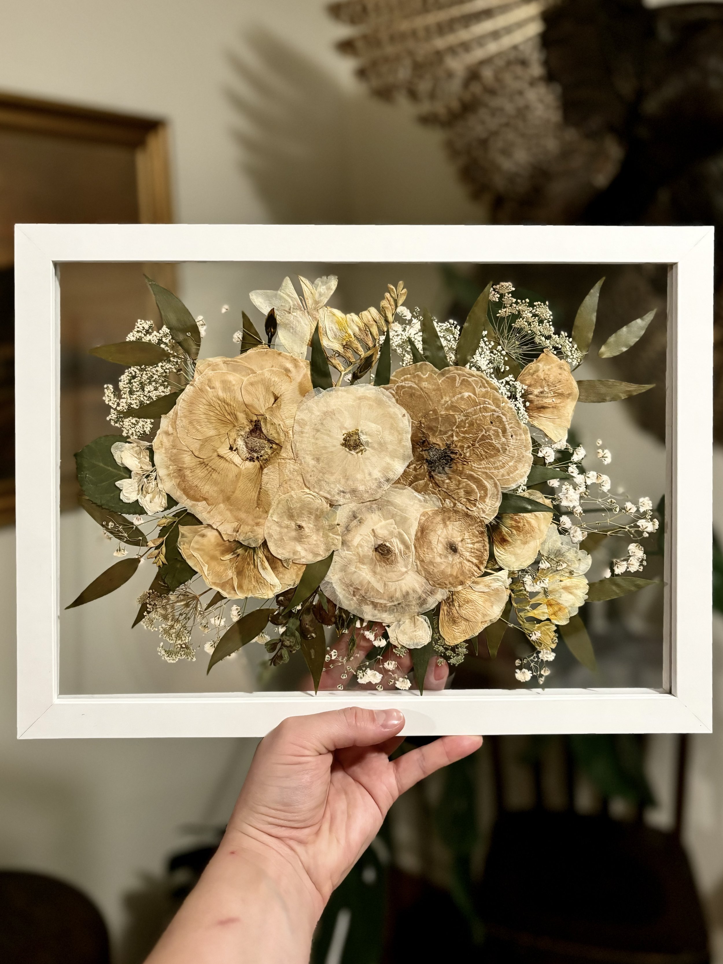 A hand holding a framed display of dried flowers and greenery. The flowers are mostly beige and brown, with small white accent flowers and green leaves.