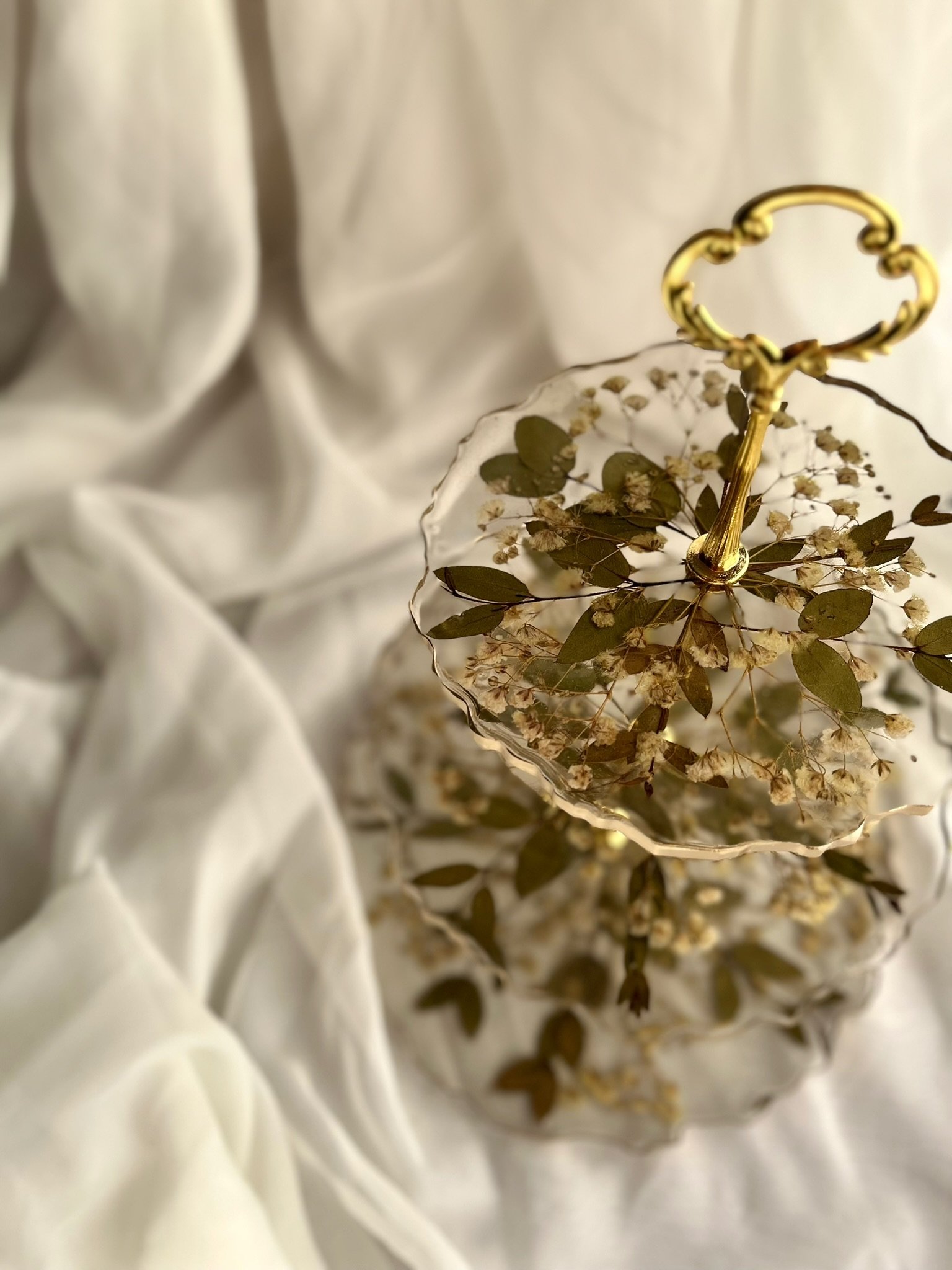A two-tiered glass and gold decorative serving stand with dried leaves and small flowers inside, set on white fabric.