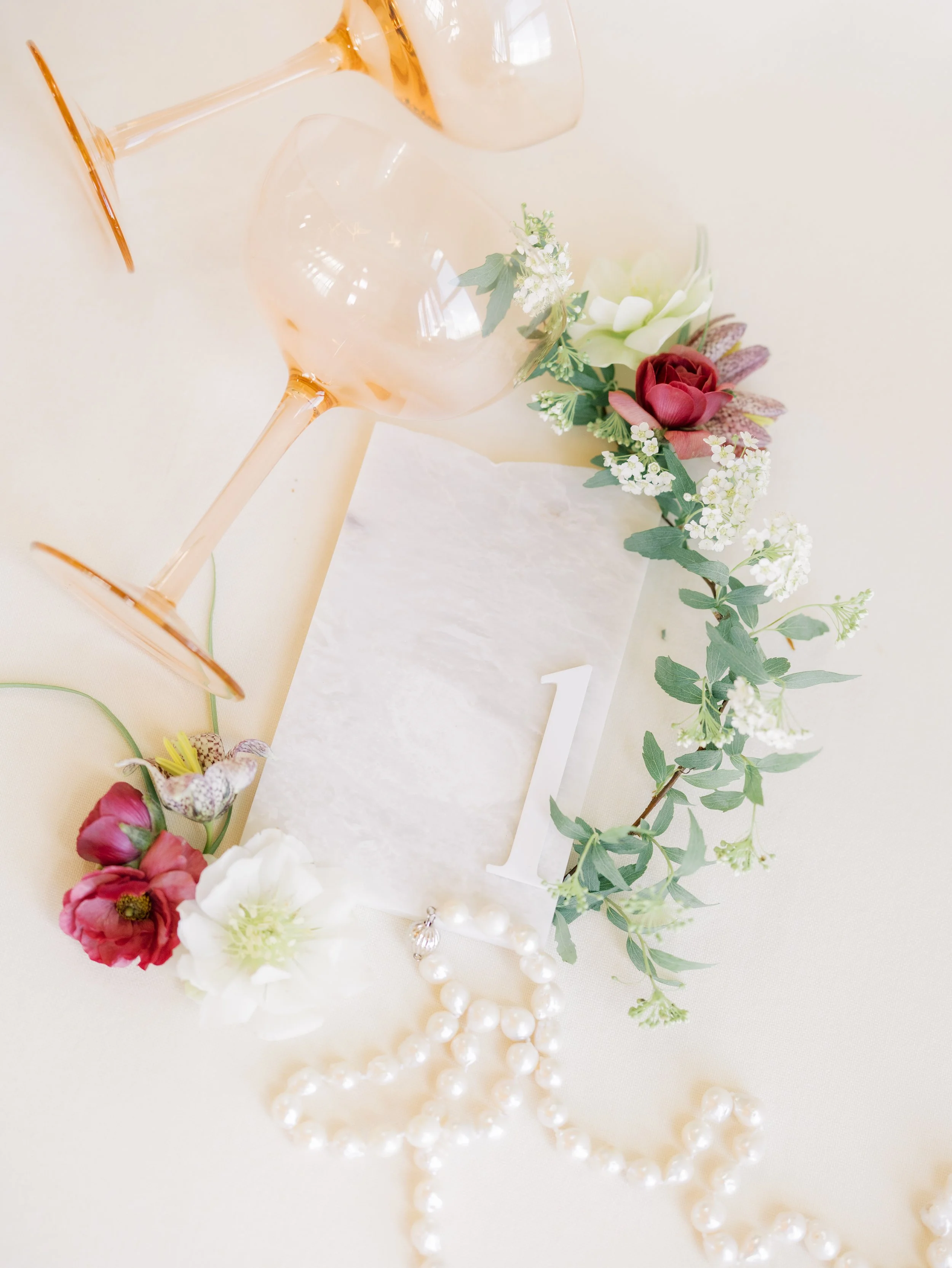 A still life arrangement with empty pink wine glasses, a white flower, pink and red flowers, eucalyptus leaves, a string of pearls, and a white card with the number 1 on a light-colored surface.
