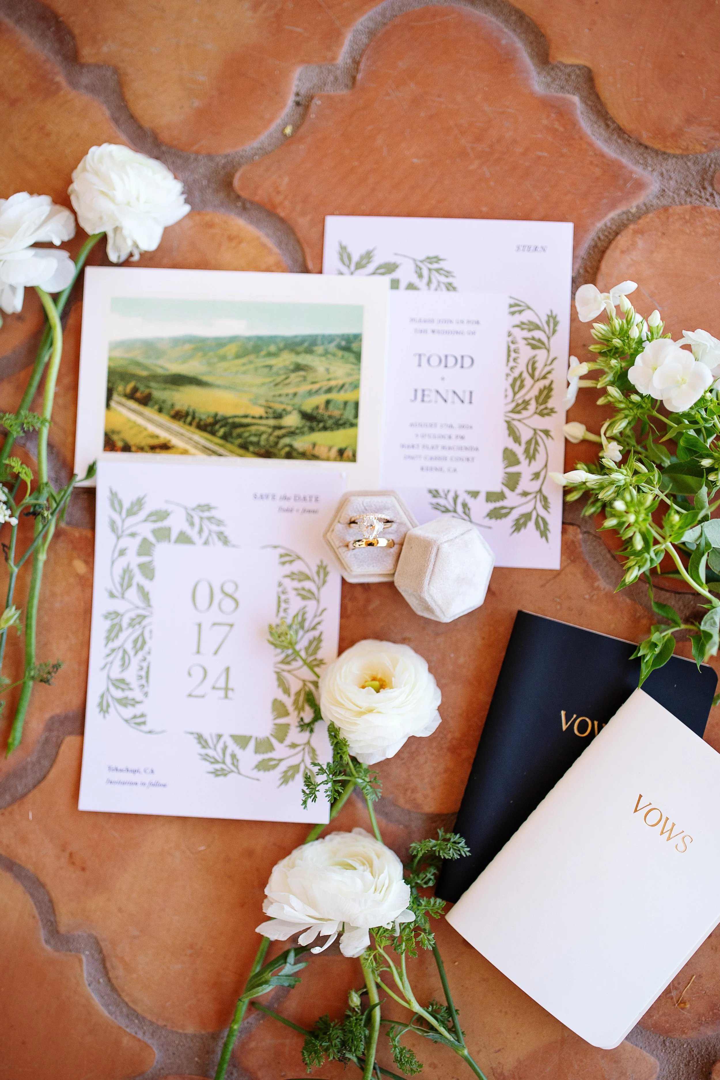 Wedding invitation suite with floral motifs and greenery, white flowers, and a small box of rings on a terracotta tile surface.