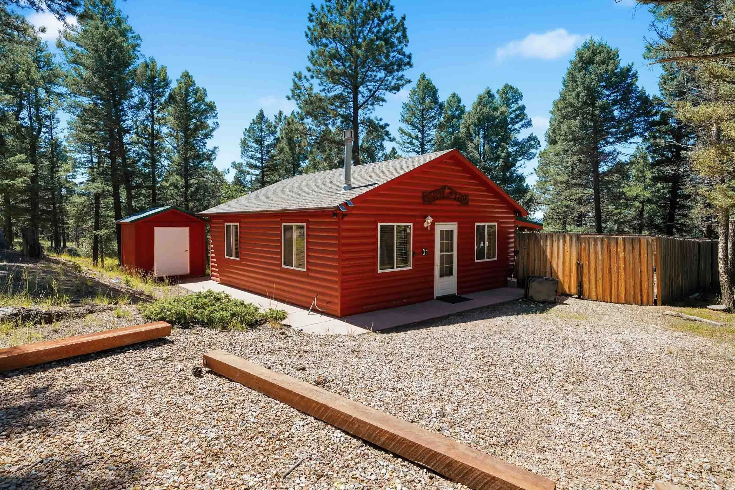 A red house with a gray roof, surrounded by tall pine trees, on a clear, sunny day, with a gravel driveway and a wooden fence on the side.