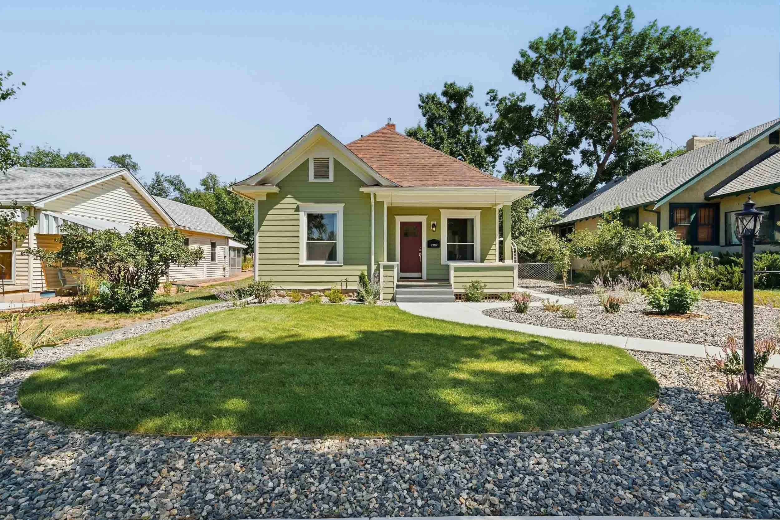 Front view of a small light green house with a brown roof, a red door, and a small porch with white railing. The house is surrounded by a well-manicured lawn, gravel paths, and landscaped garden beds, with neighboring houses visible on each side and 