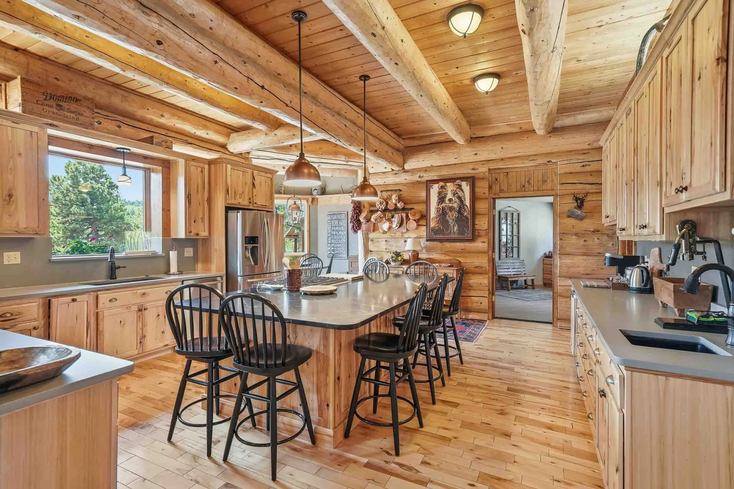 Rustic kitchen with pine wood cabinets, a central island with bar stools, stainless steel appliances, and wood-beam ceiling, overlooking a living area.