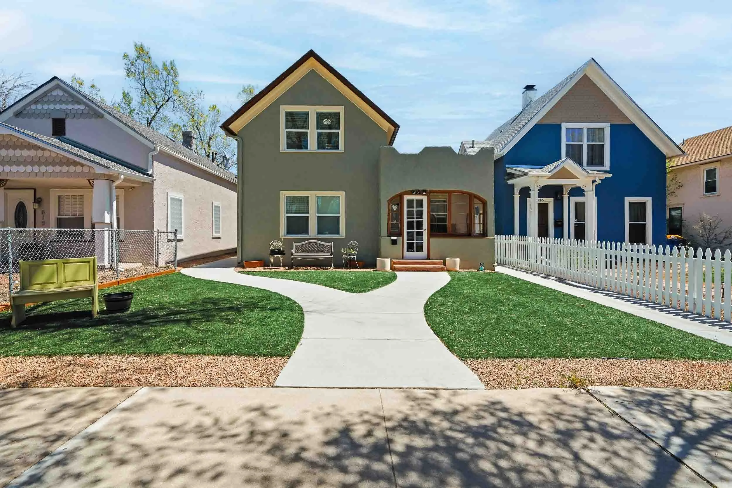 A backyard with a paved pathway, green lawn, white fence on the right, and a small house painted in gray with a porch in the center. There are two neighboring houses, one pink and one blue, visible on either side.
