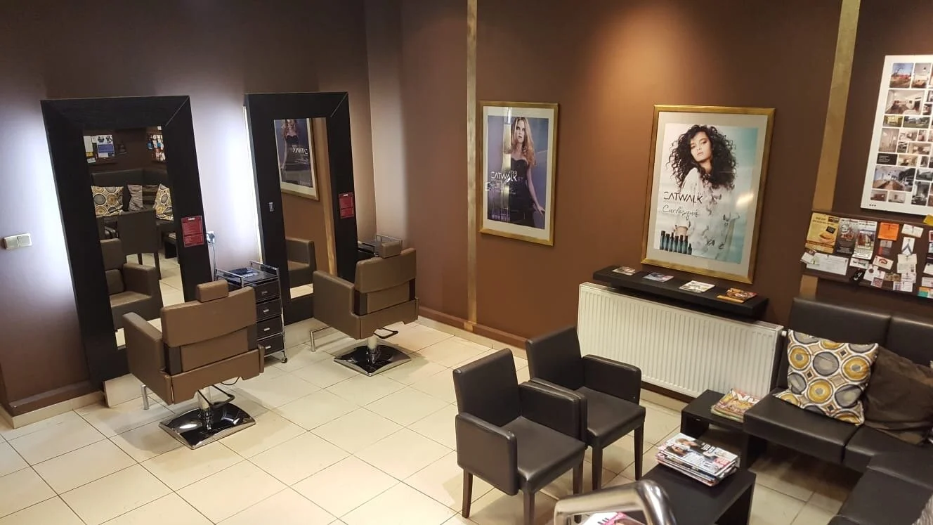 Interior of a modern hair salon with styling chairs, large mirrors, framed posters on the walls, and a waiting area with couches and magazines.