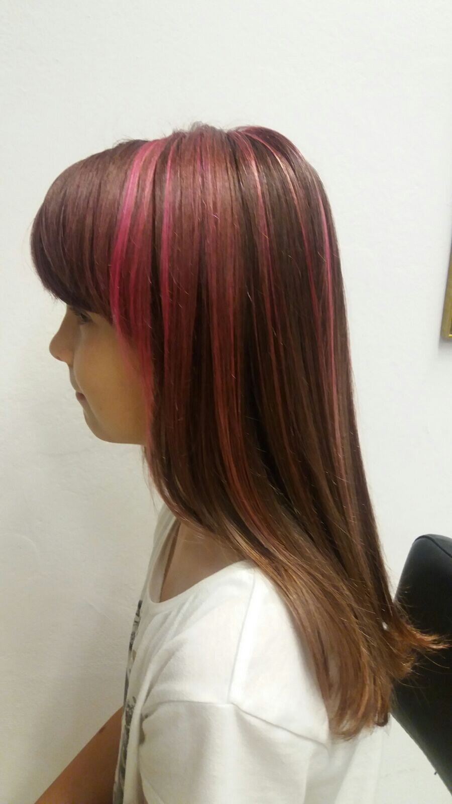 Side view of a person with straight brown hair and pink highlights, wearing a white shirt, on a plain background.