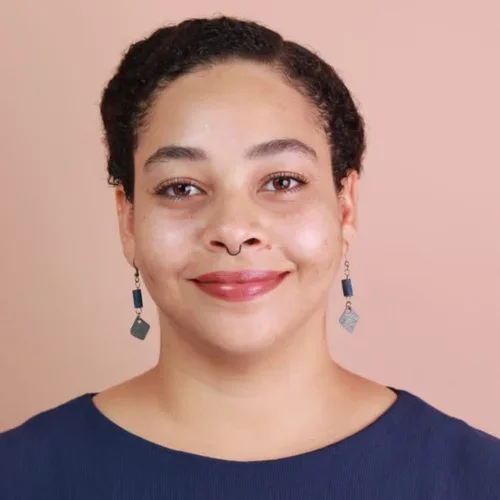 Smiling woman with dark hair, wearing a dark textured blazer, stands with arms crossed in a well-lit room.