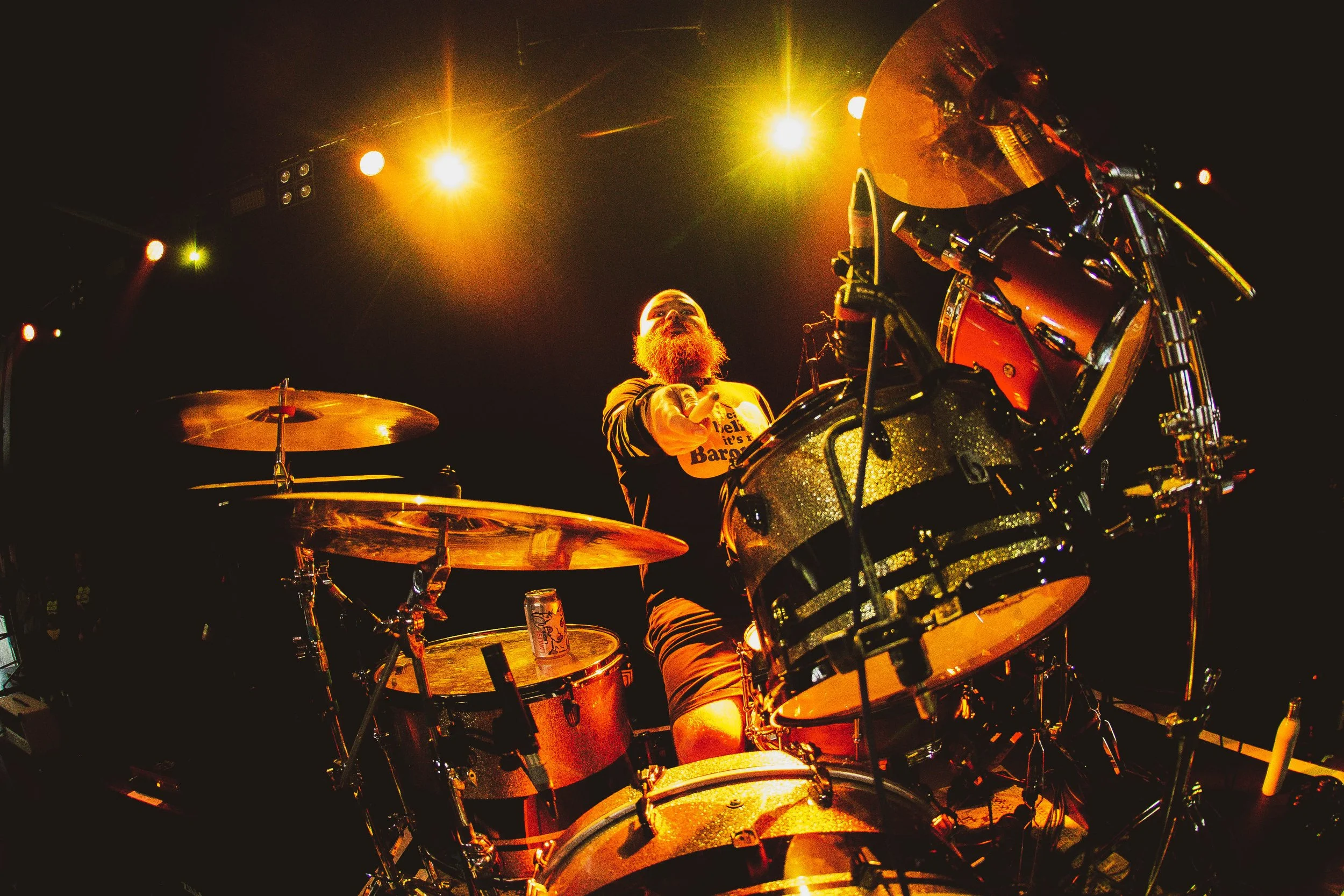 A musician with a beard playing drums on stage, illuminated by bright yellow stage lights.