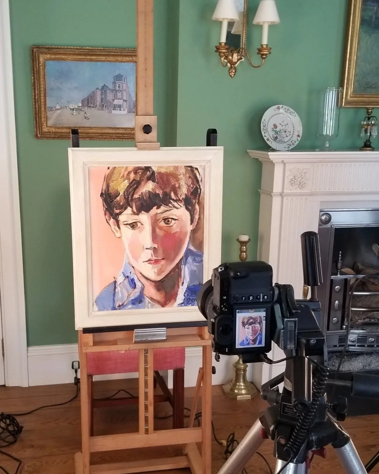 A portrait painting of a young boy on an easel in a room with a green wall, gold-frame paintings, a fireplace, and vintage decor, being photographed with a camera on a tripod.