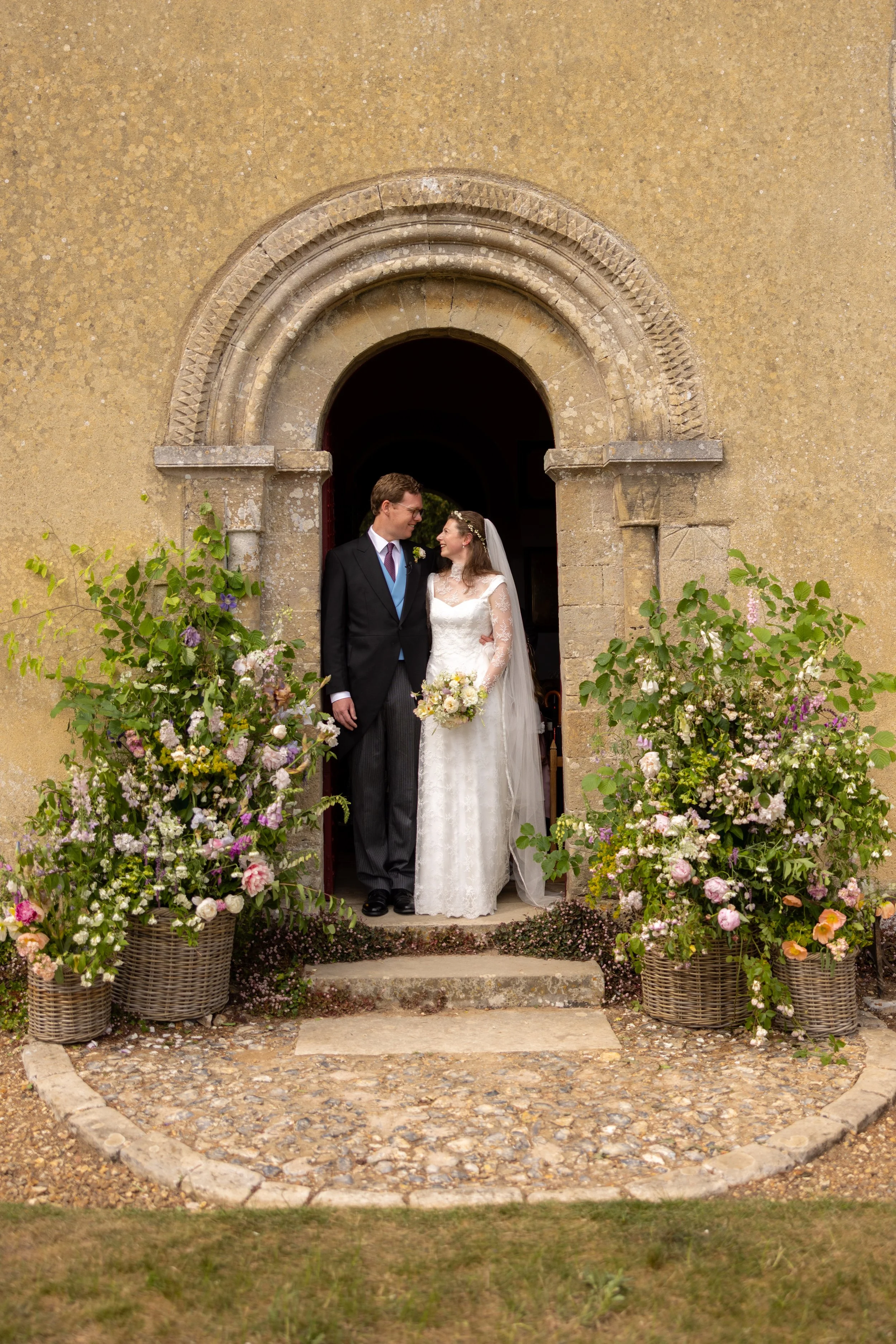 A bride and groom standing in front of a church entrance decorated with flowers, looking at each other happily.