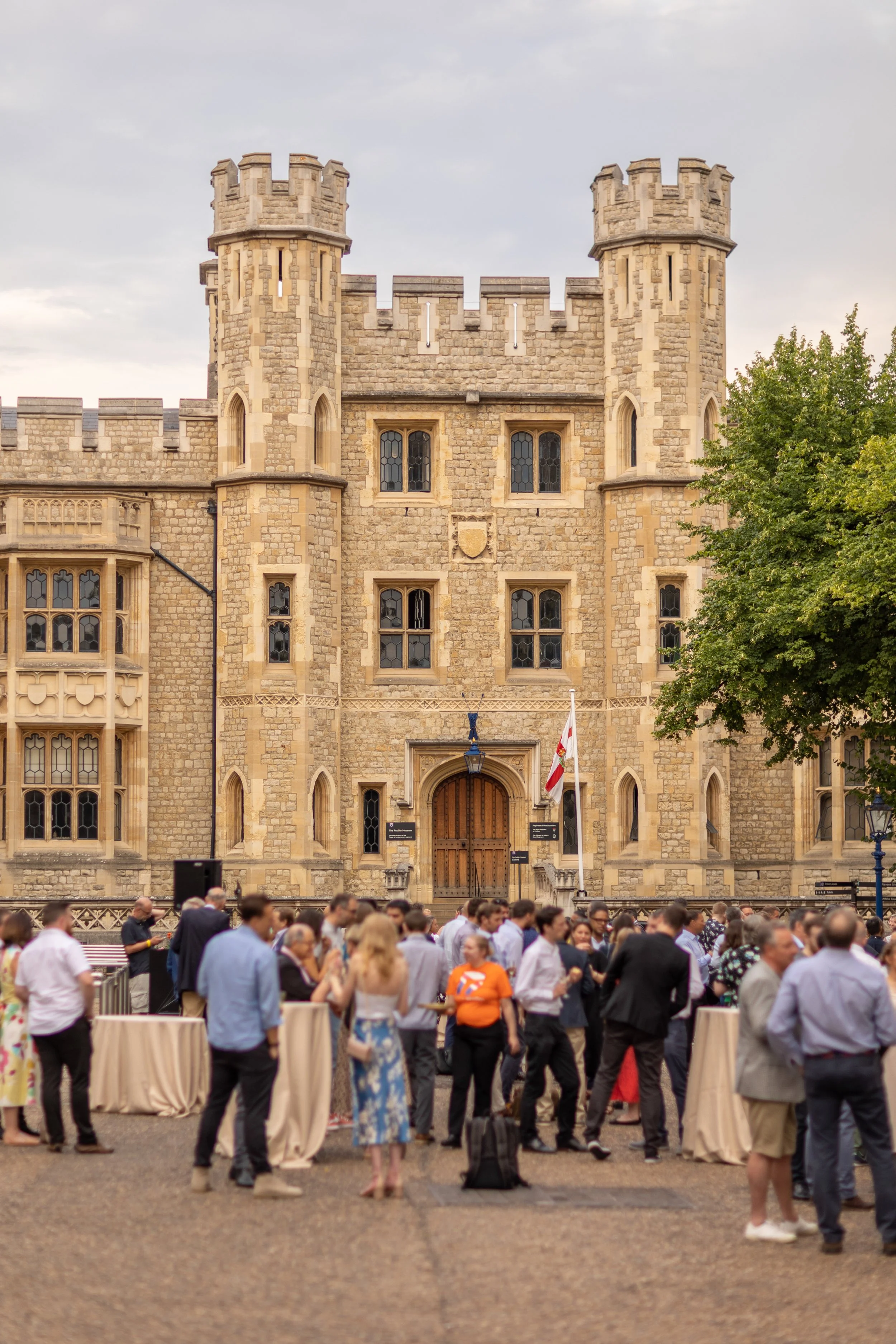 People gathered in front of a historic castle-like building with a large wooden door, stone walls, and two towers, during an outdoor event.
