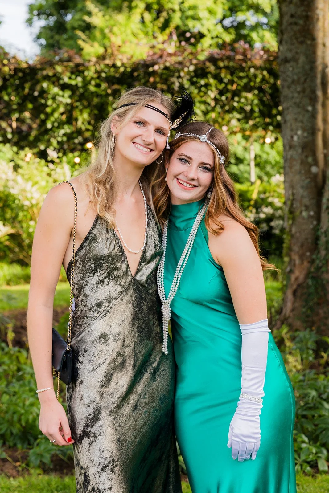 Two women smiling and dressed in elegant gowns standing outdoors with greenery and trees in the background.