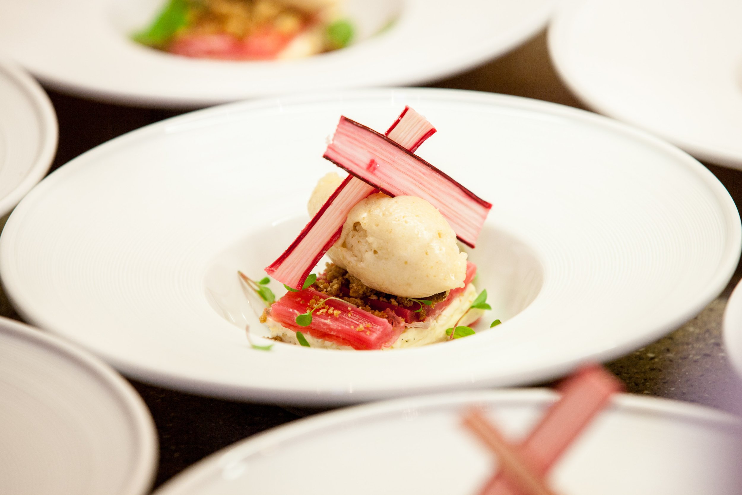 An artful plated dish featuring a scoop of white sorbet, pink and white crab sticks, green microgreens, and a bed of crumbled crumbs on a white plate.