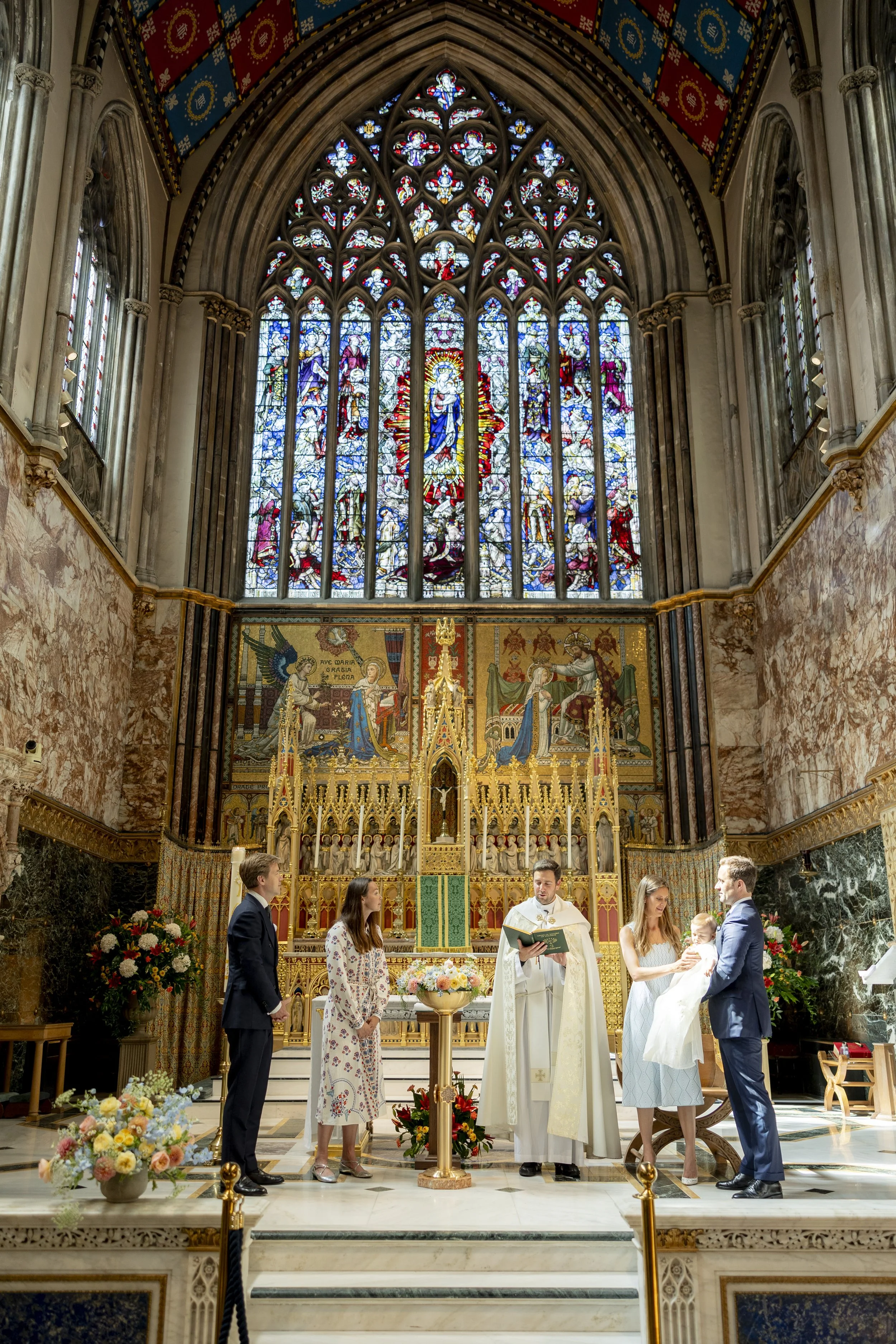 A baptism ceremony taking place inside a grand church with stained glass windows, a priest, a baby, and family members.