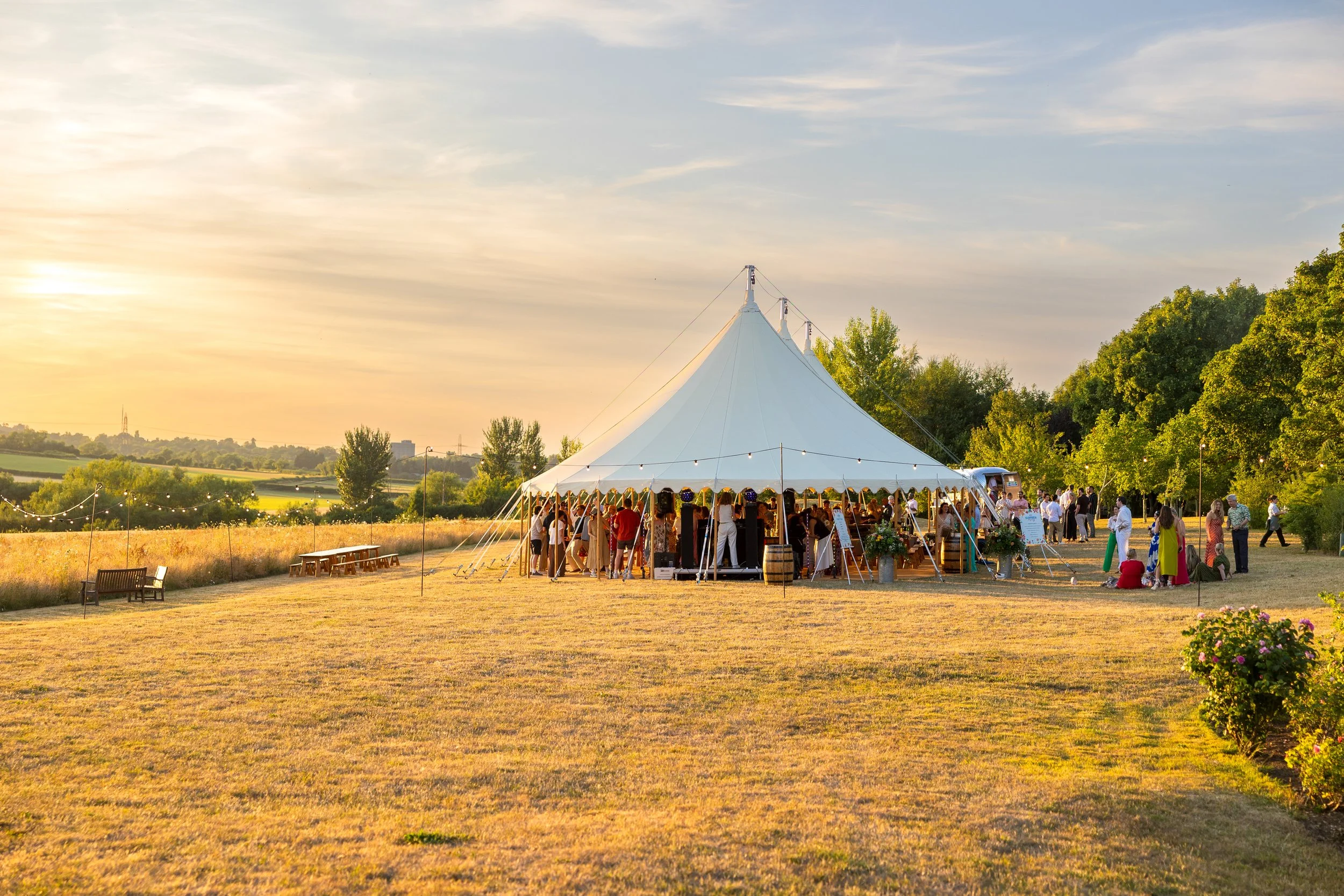 A large white event tent set up outdoors during sunset with many people gathered underneath, celebrating on a grassy field surrounded by trees and open landscape.