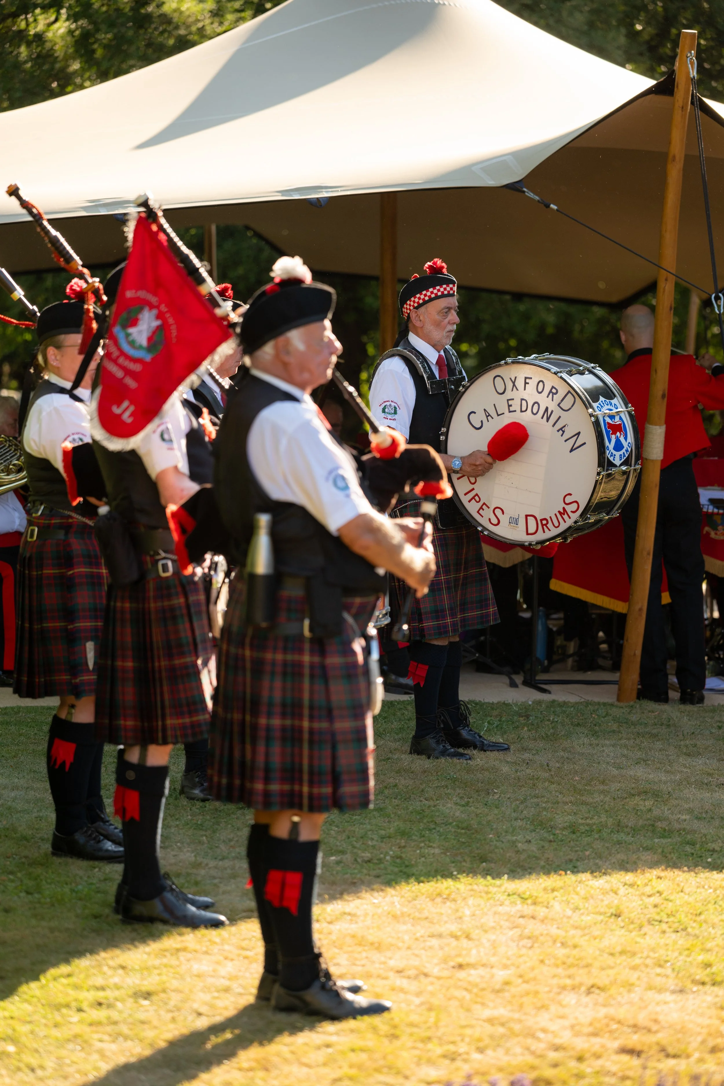 A Scottish pipe band performing outdoors under a large tent, dressed in traditional kilts and uniforms, with one member playing a bass drum labeled "Oxford Caledonian Pipes and Drums."