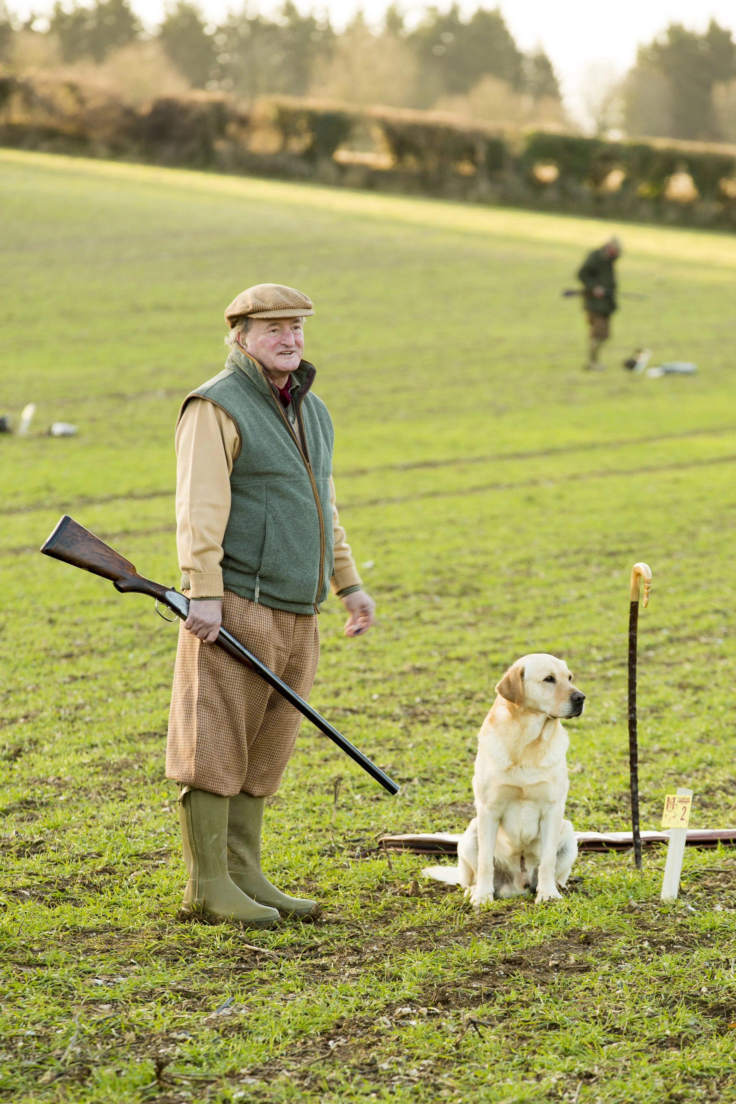 An elderly man holding a shotgun sitting next to a yellow labrador dog in a grassy field with a walking stick. In the background, a person is seen with hunting gear. The setting appears to be a hunting or outdoor activity area.