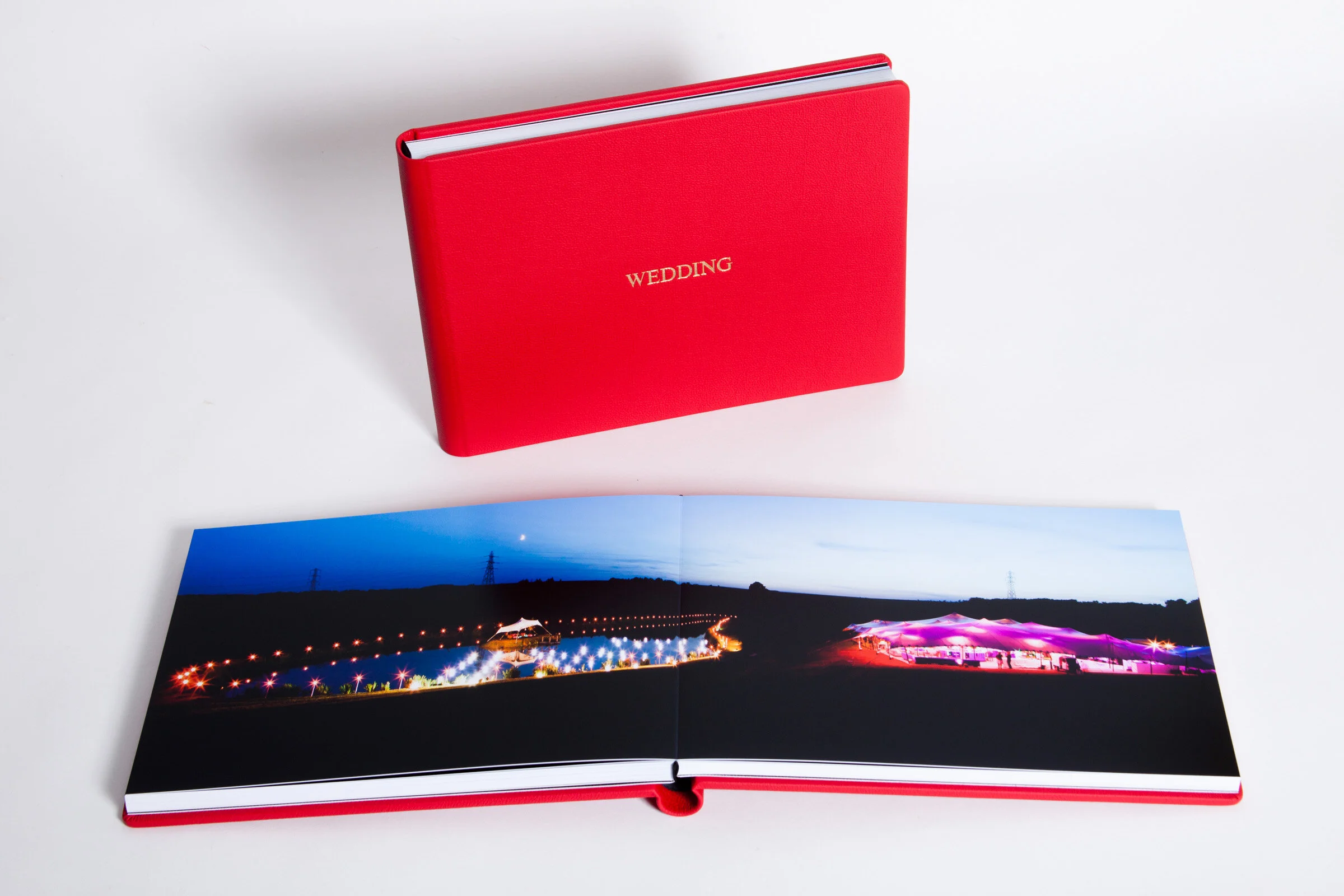 A red wedding photo album with the word 'WEDDING' in gold on the cover. The album is open showing a night scene with colorful lights and a small white tent.