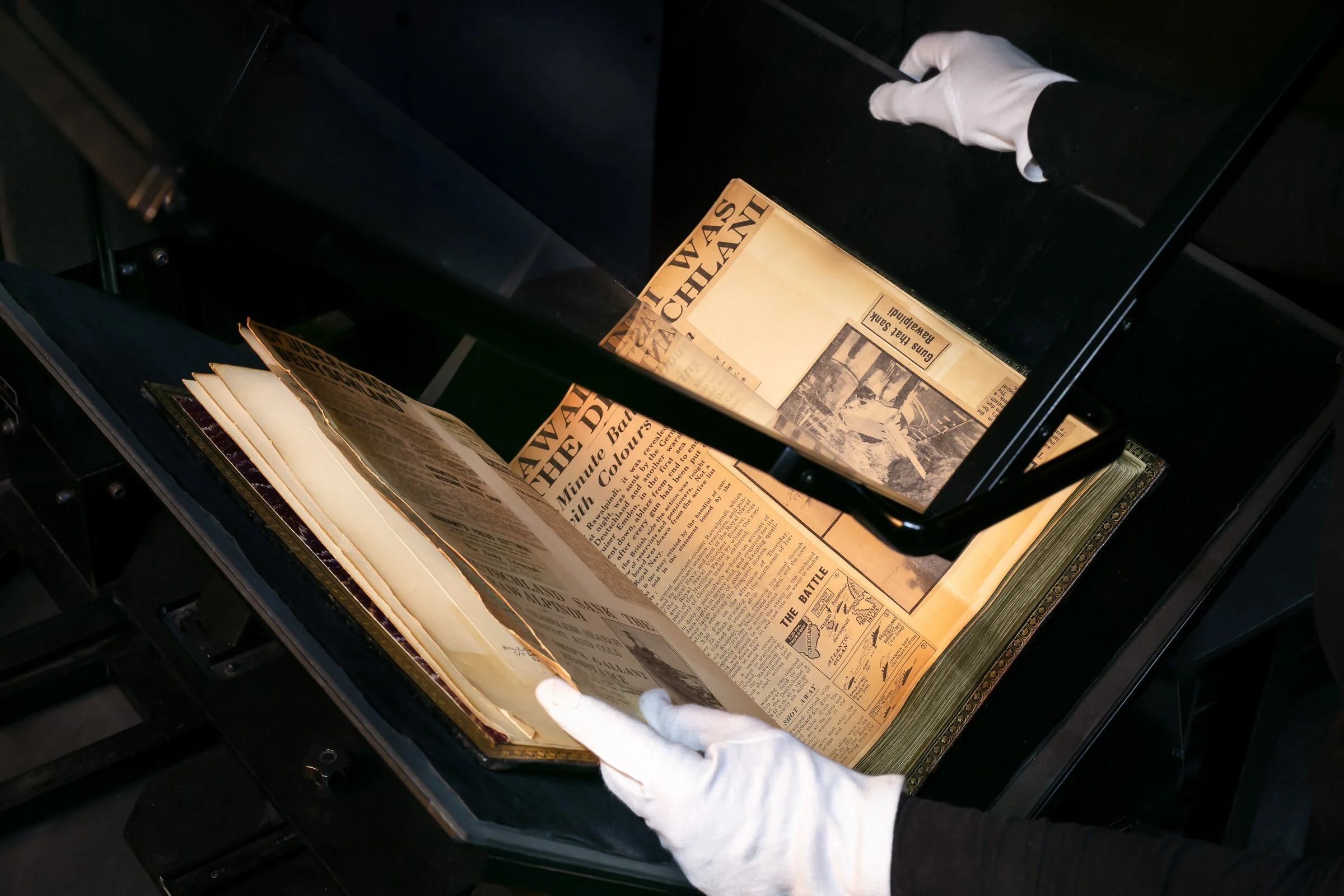 A person in white gloves handling an old newspaper or publication in a display case or preservation setting.