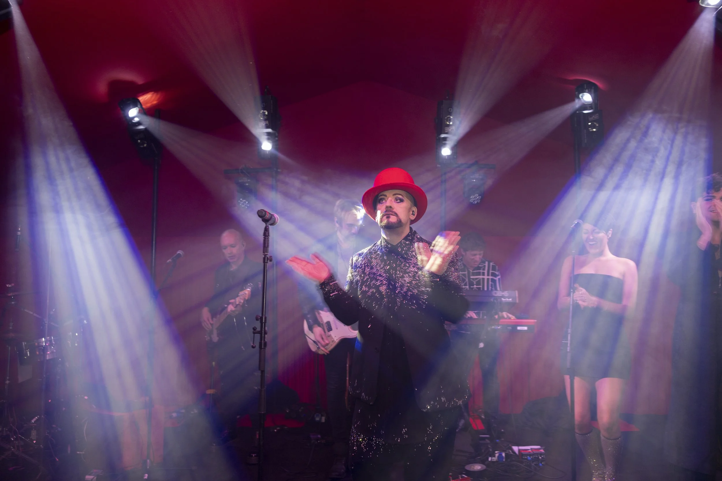 A performer in a shiny black outfit and a red hat on stage, clapping with a band playing instruments and other performers in the background, under colorful stage lighting.