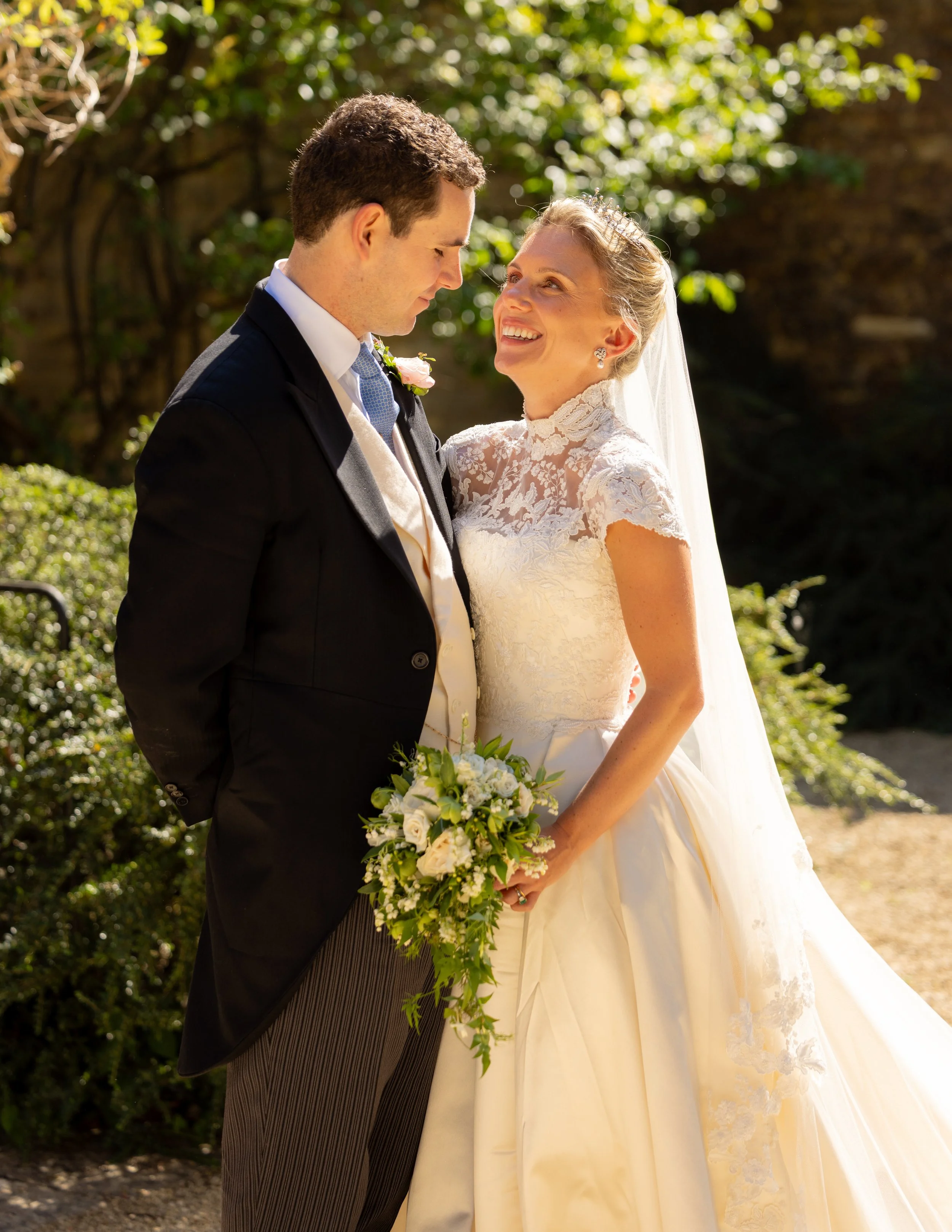 A bride and groom are standing close together, smiling at each other outdoors during the daytime. The bride is wearing a lace wedding dress and a veil, holding a bouquet of white and green flowers. The groom is dressed in a black tuxedo with a light-