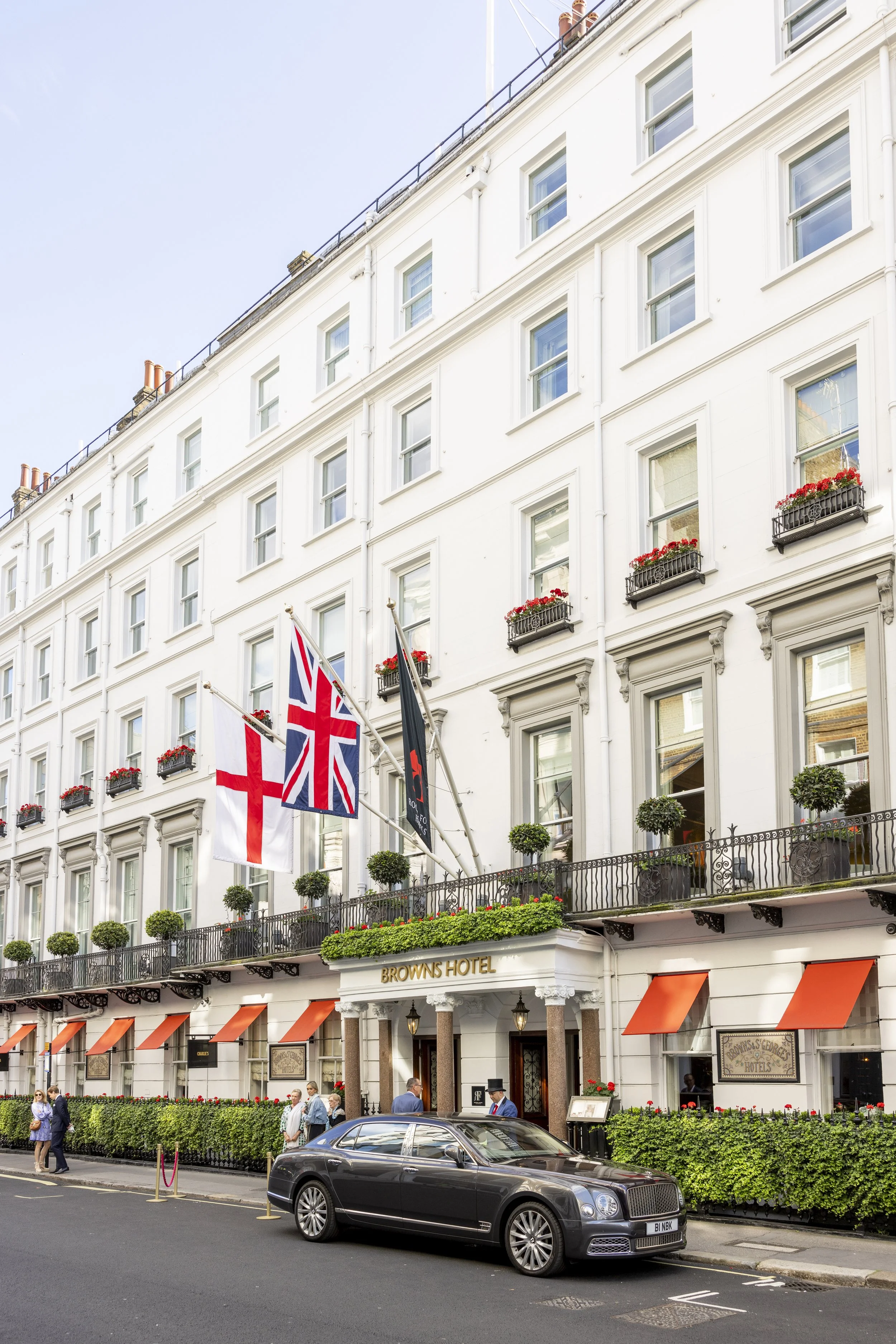 Exterior view of Browns Hotel, a white multi-story building with 4 flags - England, the UK, and another. The hotel has potted plants and flower boxes on the balconies, with people walking in front and a black luxury car parked on the street.