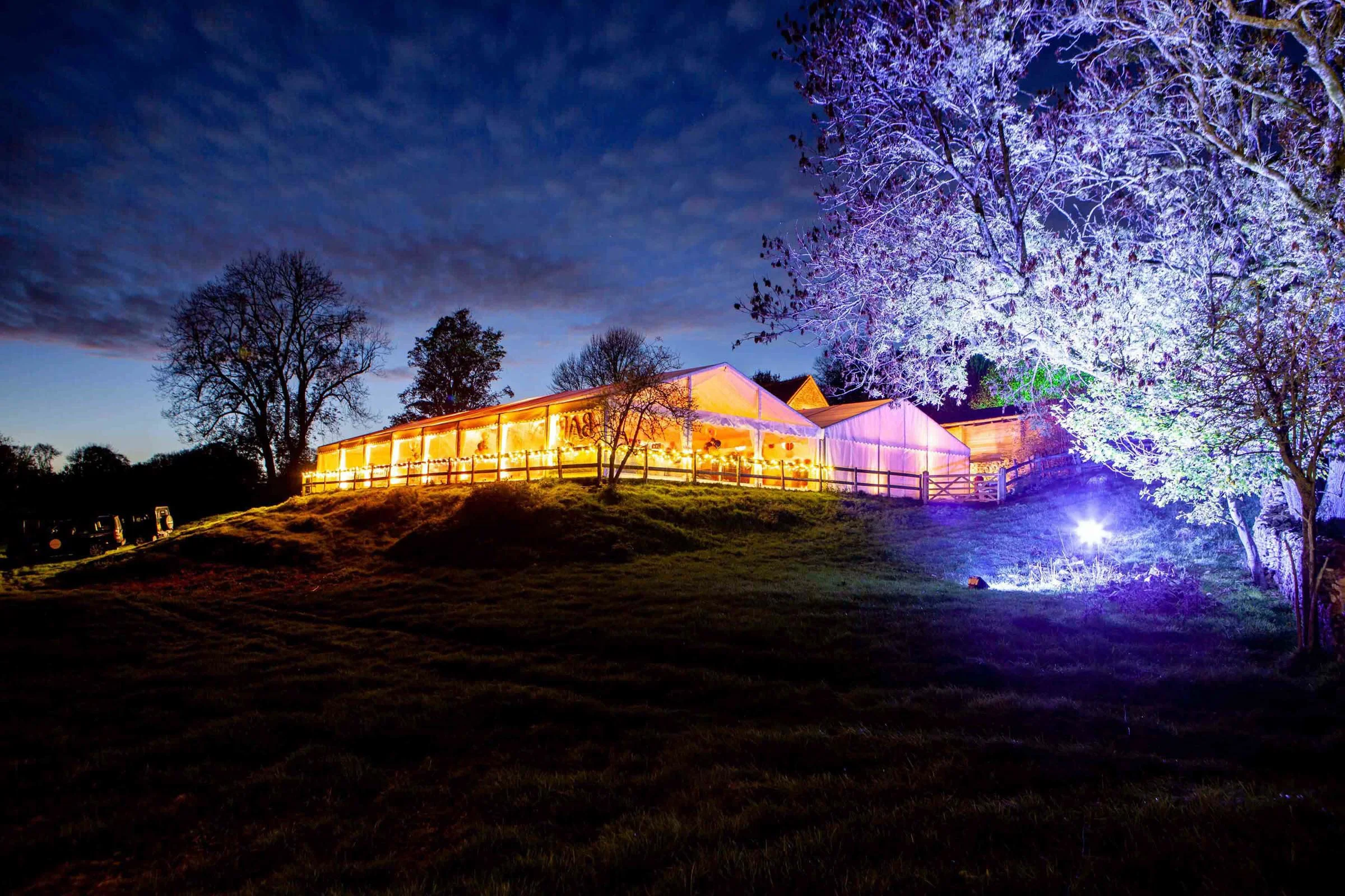 A brightly lit event tent on a grassy hill during nighttime, with colorful lighting illuminating surrounding trees and the dark sky in the background.