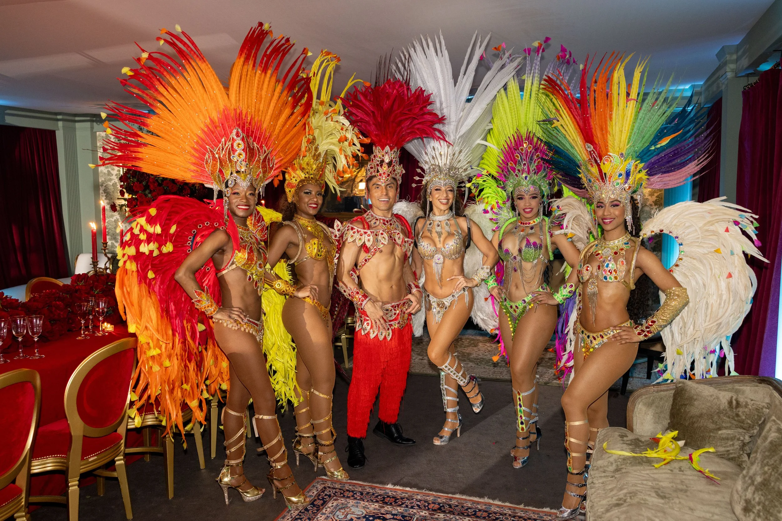 Group of performers dressed in colorful Carnival costumes with large feathered headdresses, posing together indoors.