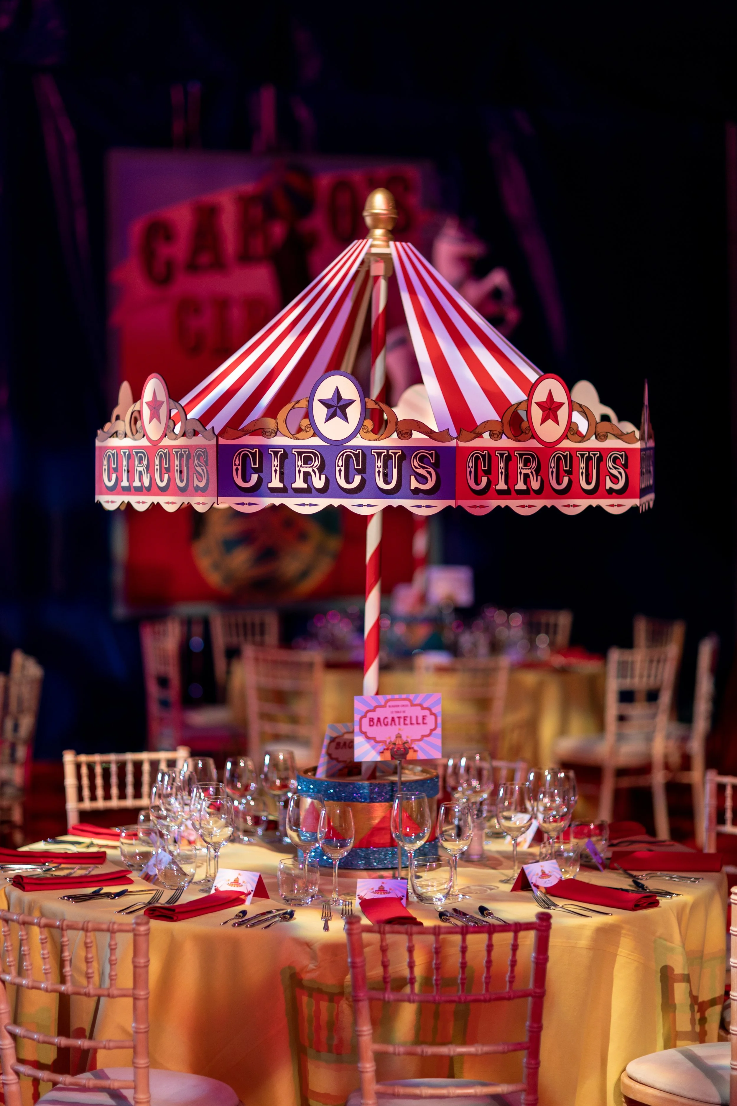 Decorative circus themed table setting with a carousel centerpiece and a round table covered with a yellow tablecloth, surrounded by wooden chairs, set with glassware, silverware, and red napkins.