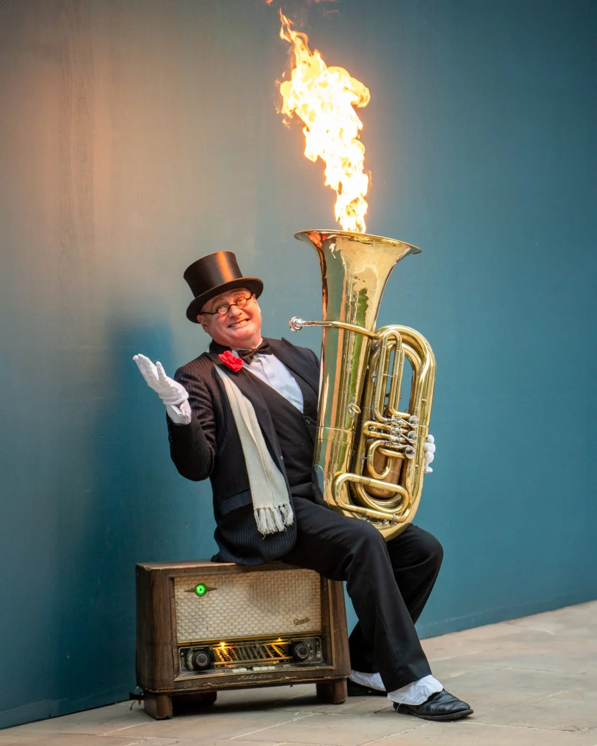 A man dressed as a circus performer, wearing a top hat, glasses, and a tuxedo with a bow tie and flower, is sitting on a vintage radio and holding a shiny musical instrument, possibly a tuba. The instrument's bell is on fire, with flames shooting upw