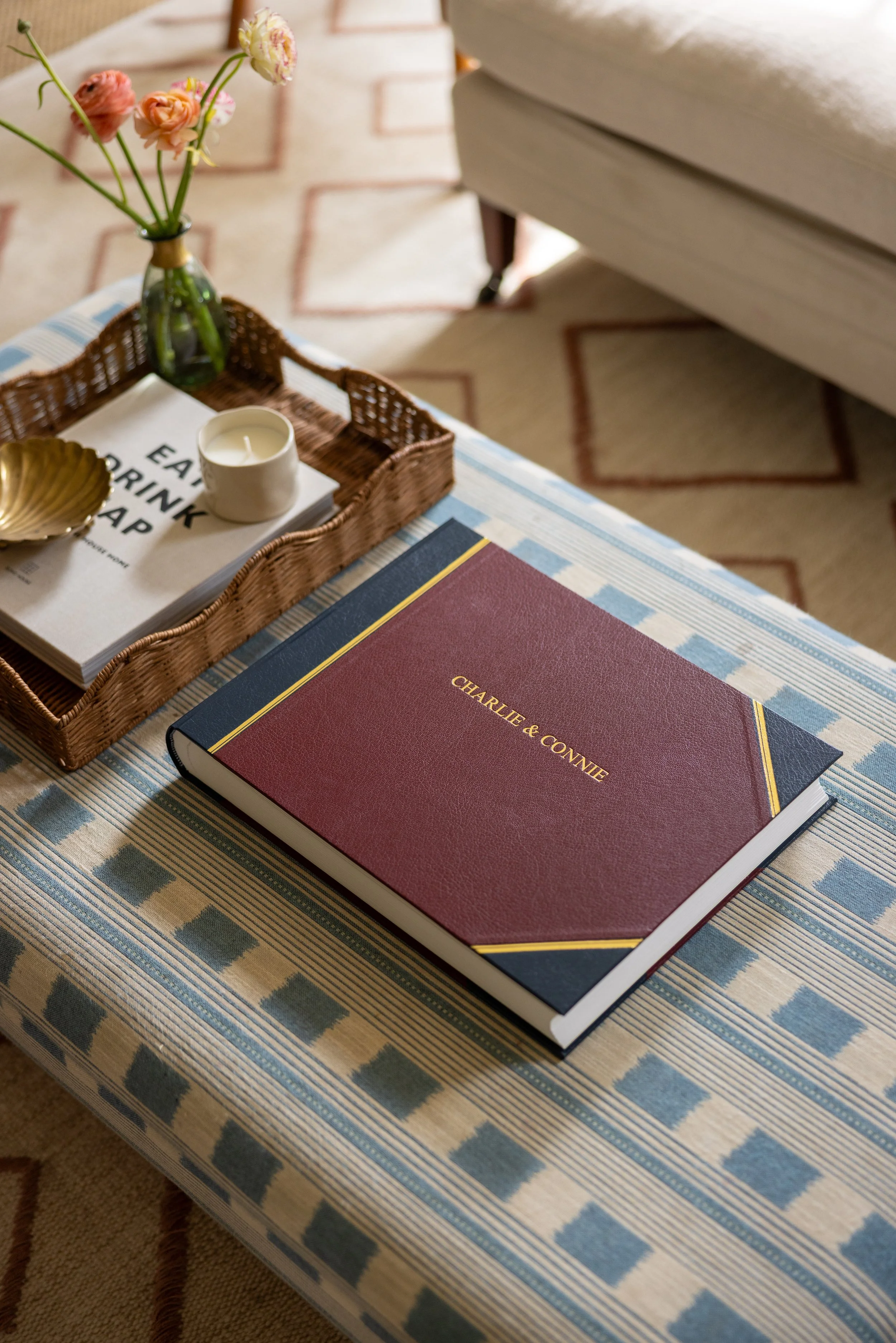 A table with a striped blue and beige tablecloth, holding a burgundy hardcover book labeled 'Charlie & Connie,' a tray with a lit white candle, a glass vase with pink and peach flowers, and a gold decorative bowl. In the background, part of a cream-c